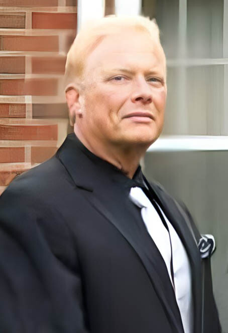 Man in a black suit with a white tie and pocket square standing against a brick wall, looking at the camera.