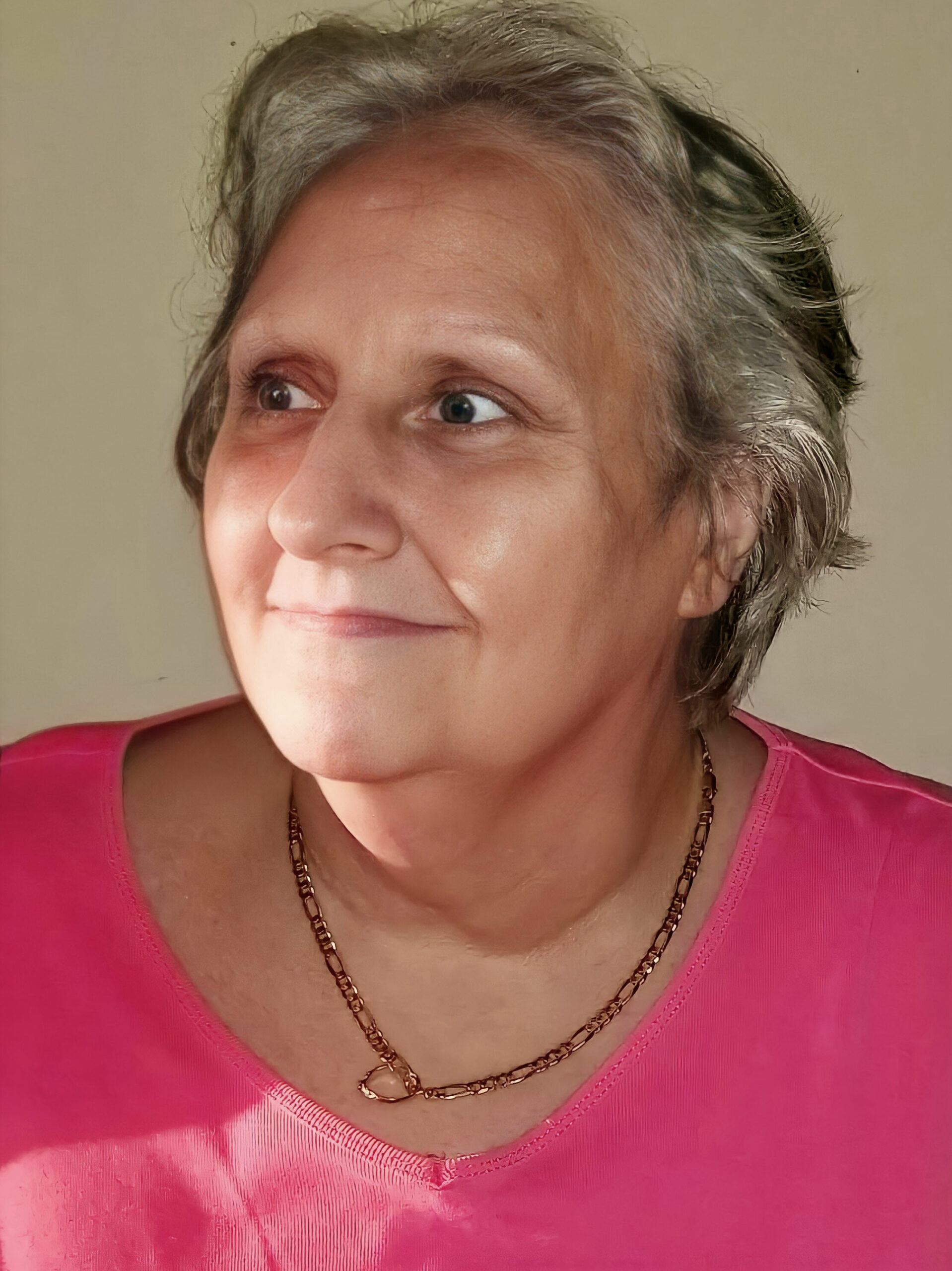 Close-up of an older woman with short gray hair, wearing a pink top and a gold necklace, looking to the right against a neutral background.