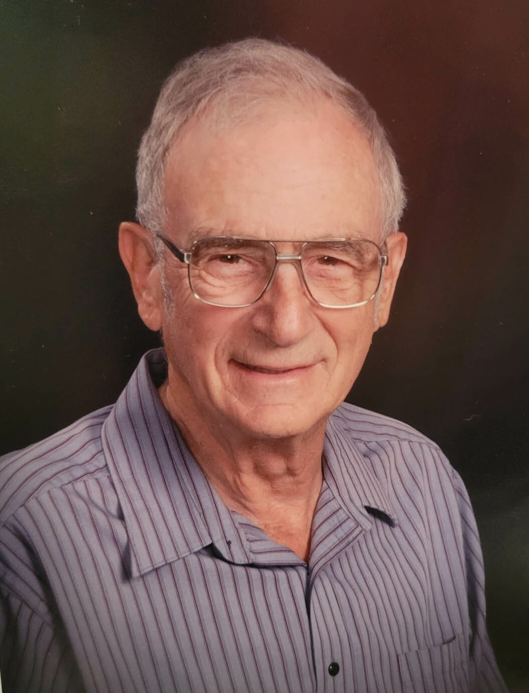 Older man with short white hair and glasses, wearing a striped button-down shirt, smiling at the camera against a dark backdrop.