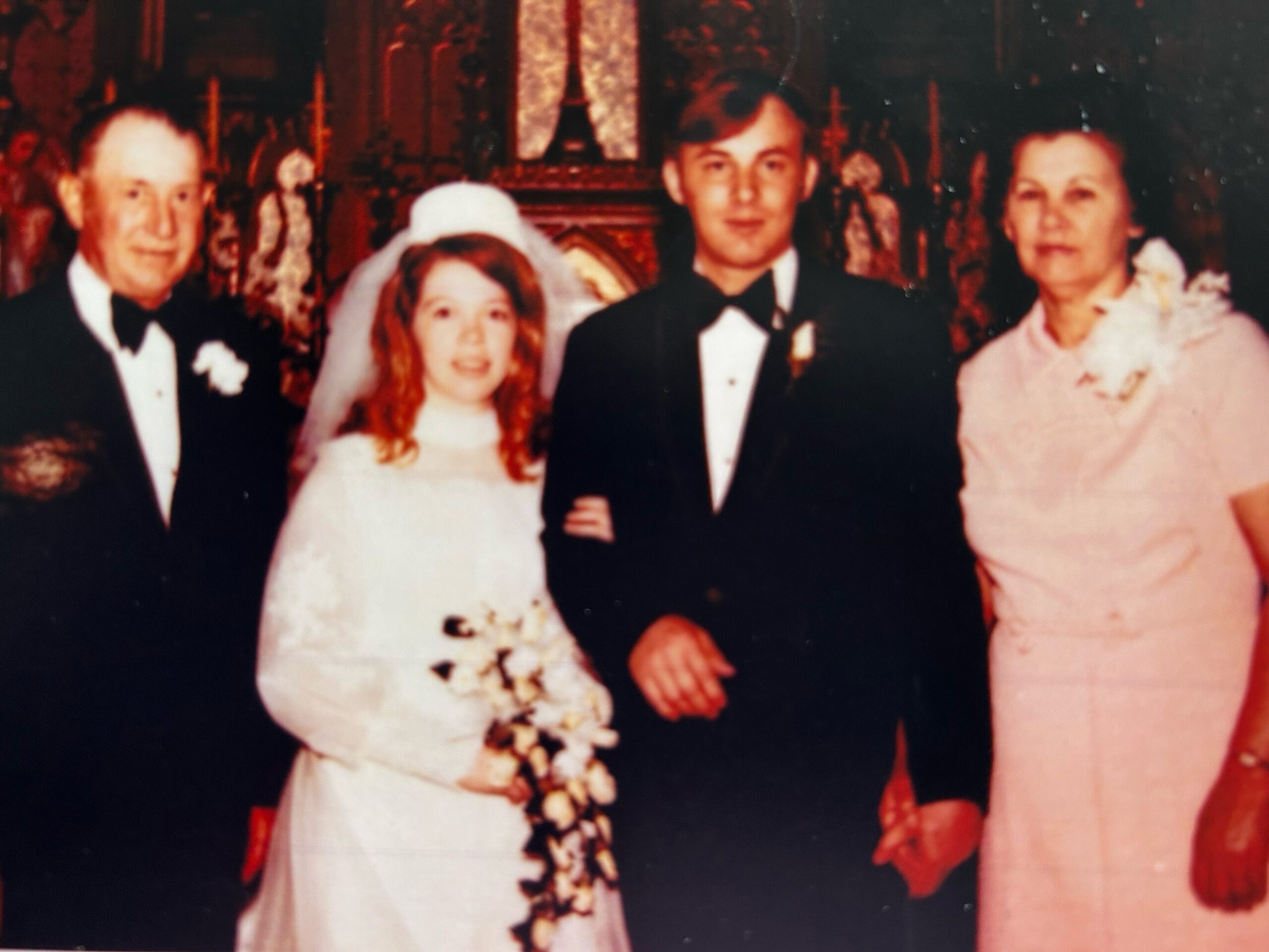 Bride in a white gown holding a cascading bouquet, standing with a groom in a black tuxedo; two other adults on either side in formal wear at a wedding photo.