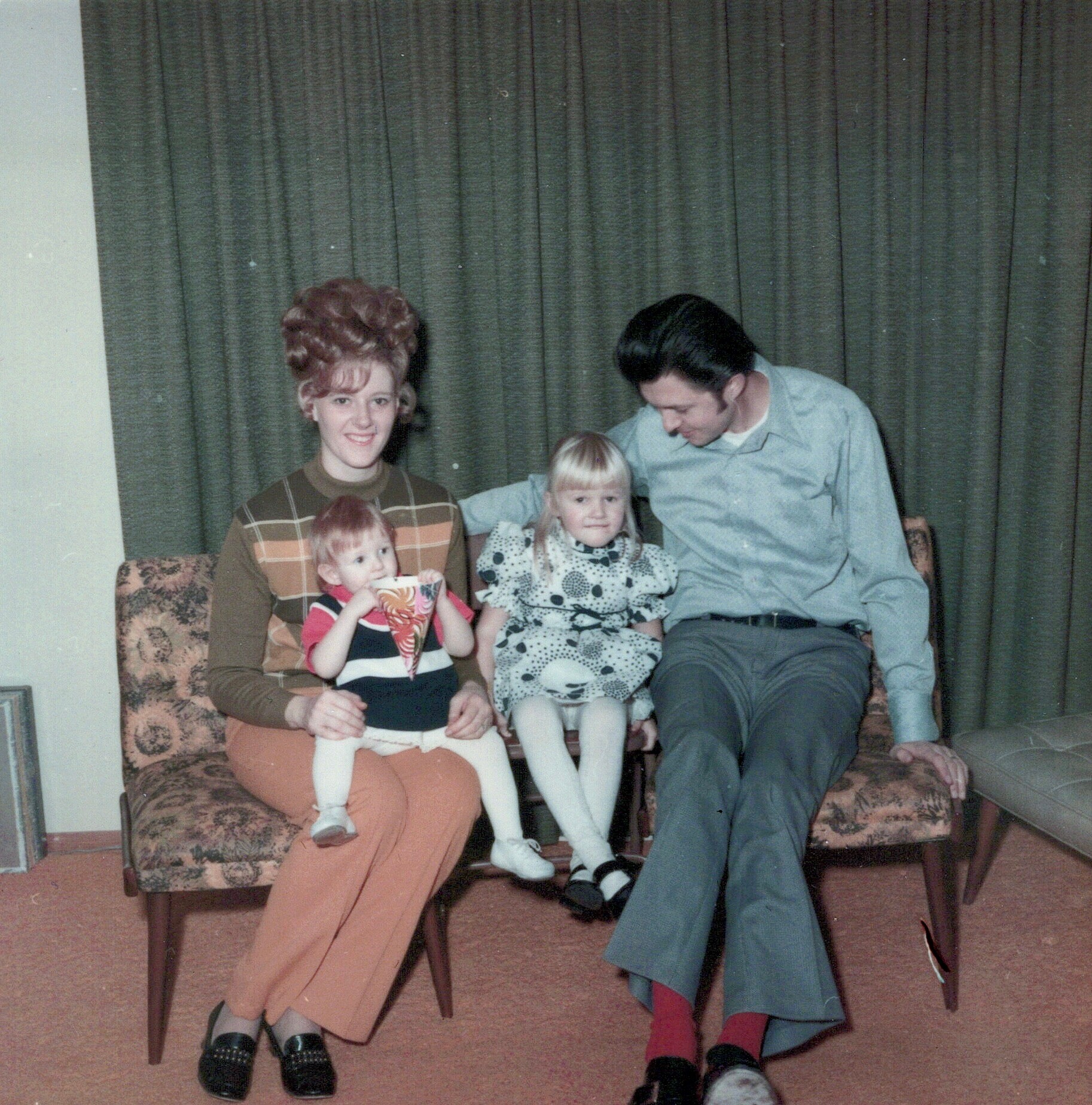Family of four posing together on a floral sofa in a living room with curtains in the background (mom, dad, two young children).
