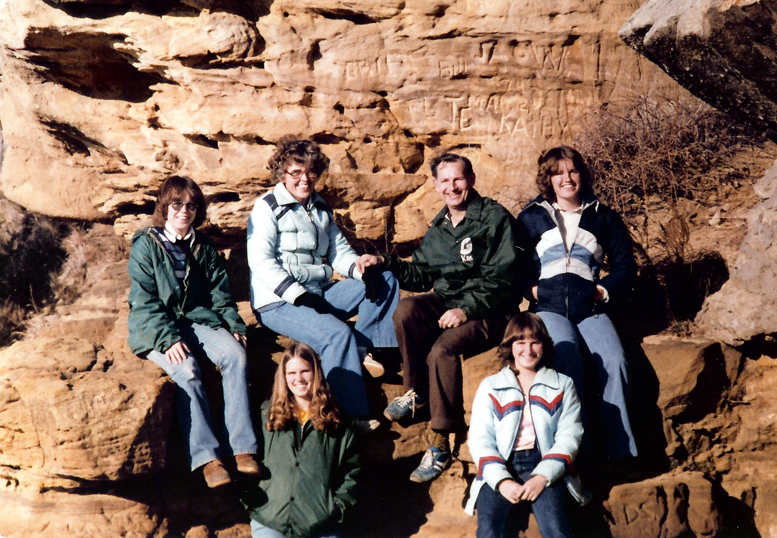 Group of people sitting on a rocky canyon wall, posing for a photo in casual jackets and jeans during daytime.
