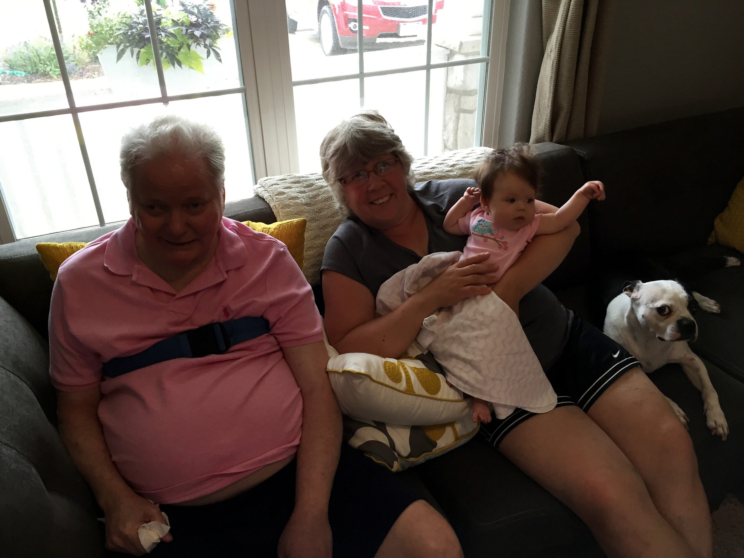 Two elderly women sit on a sofa with a baby, while a small dog rests on the right; a bright window and plants are behind them in a living room setting.