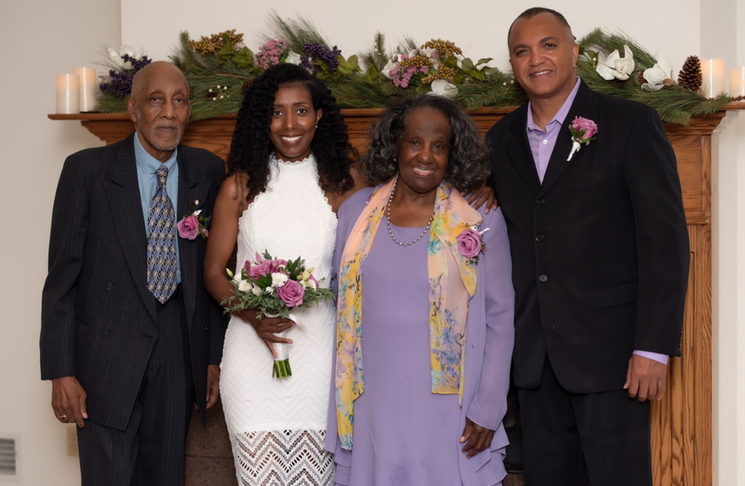 Four adults posing together at a celebration; the bride in a white lace dress holds a pink bouquet, flanked by two guests in suits and lavender outfits.