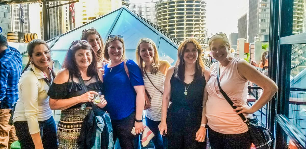 Group of seven women smiling for a photo on an outdoor terrace with a glass pyramid structure and city skyline behind them.