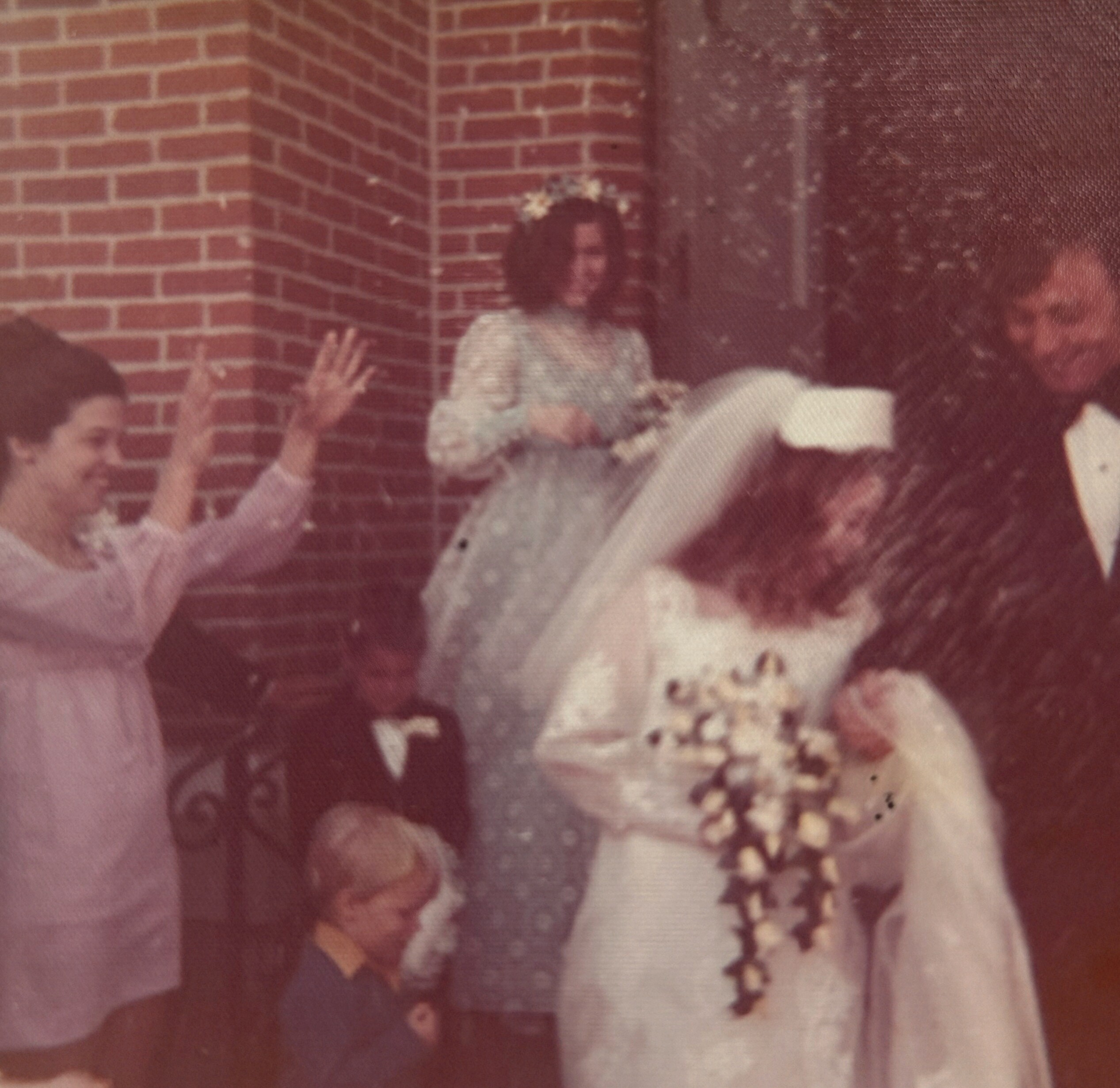 Blurry vintage photo of people gathered indoors near a brick wall, with a raised hand visible in the foreground.