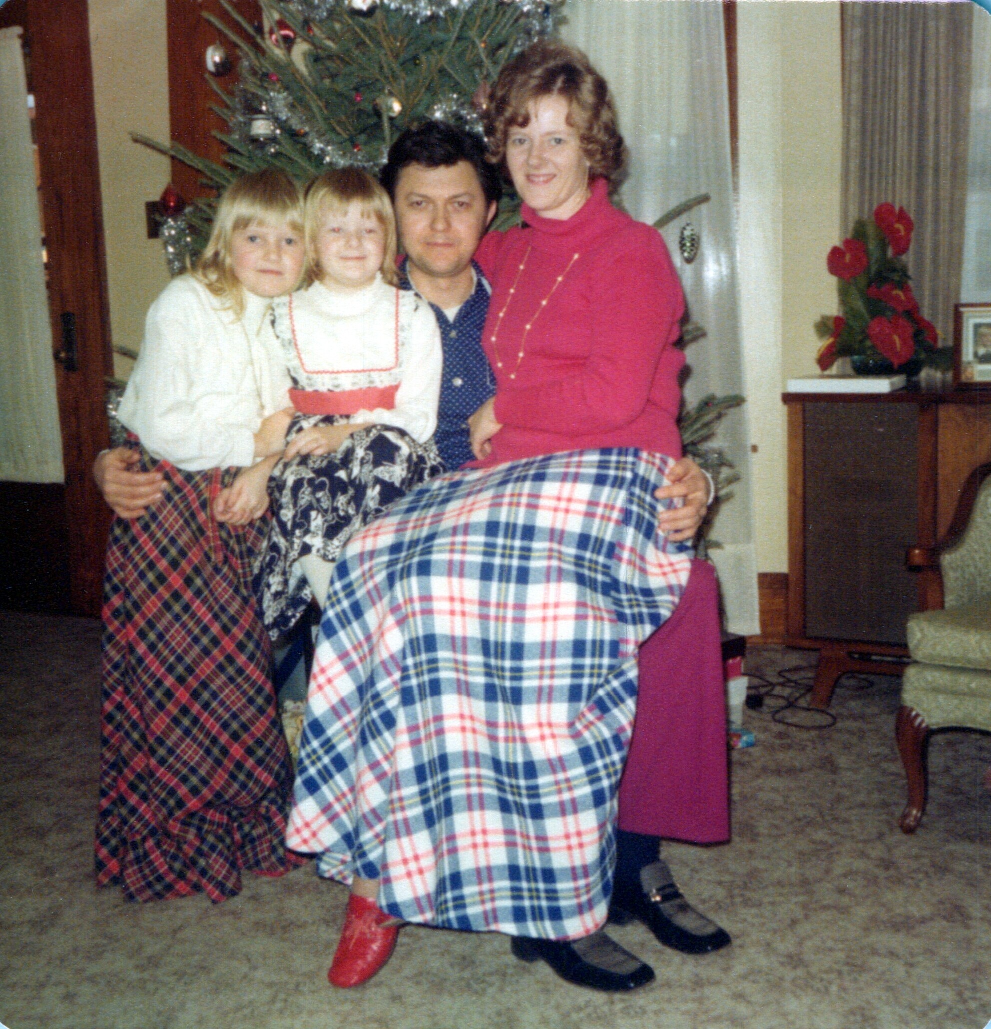 A family posing in front of a Christmas tree, with two young girls in plaid skirts and red boots, flanked by adults in a living room.