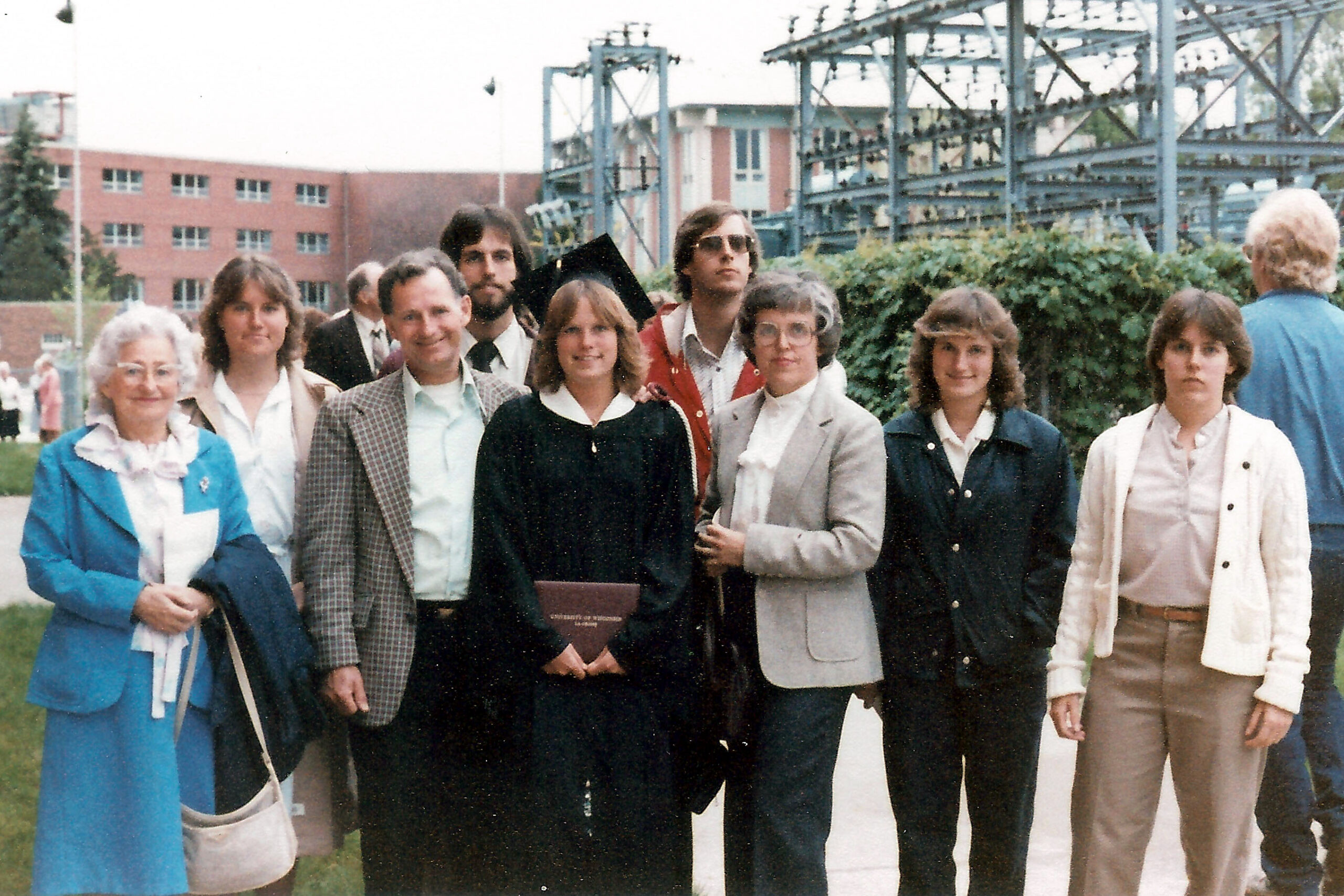 Group of people posing outdoors on a campus lawn, with one graduate in a black gown holding a diploma or folder in front.