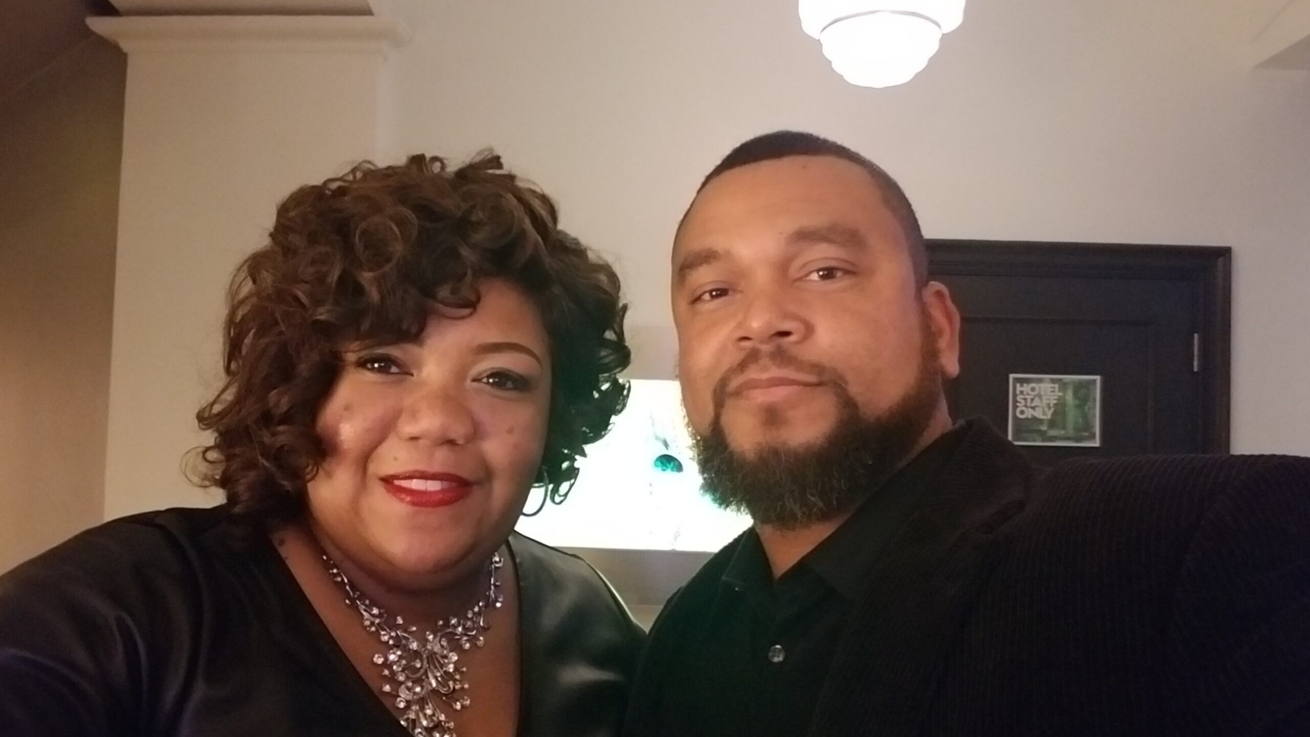 Close-up selfie of a smiling couple indoors: woman with curly hair and red lipstick on the left, man with a beard on the right, hotel hallway in background with a 'Hotel Staff Only' door sign.