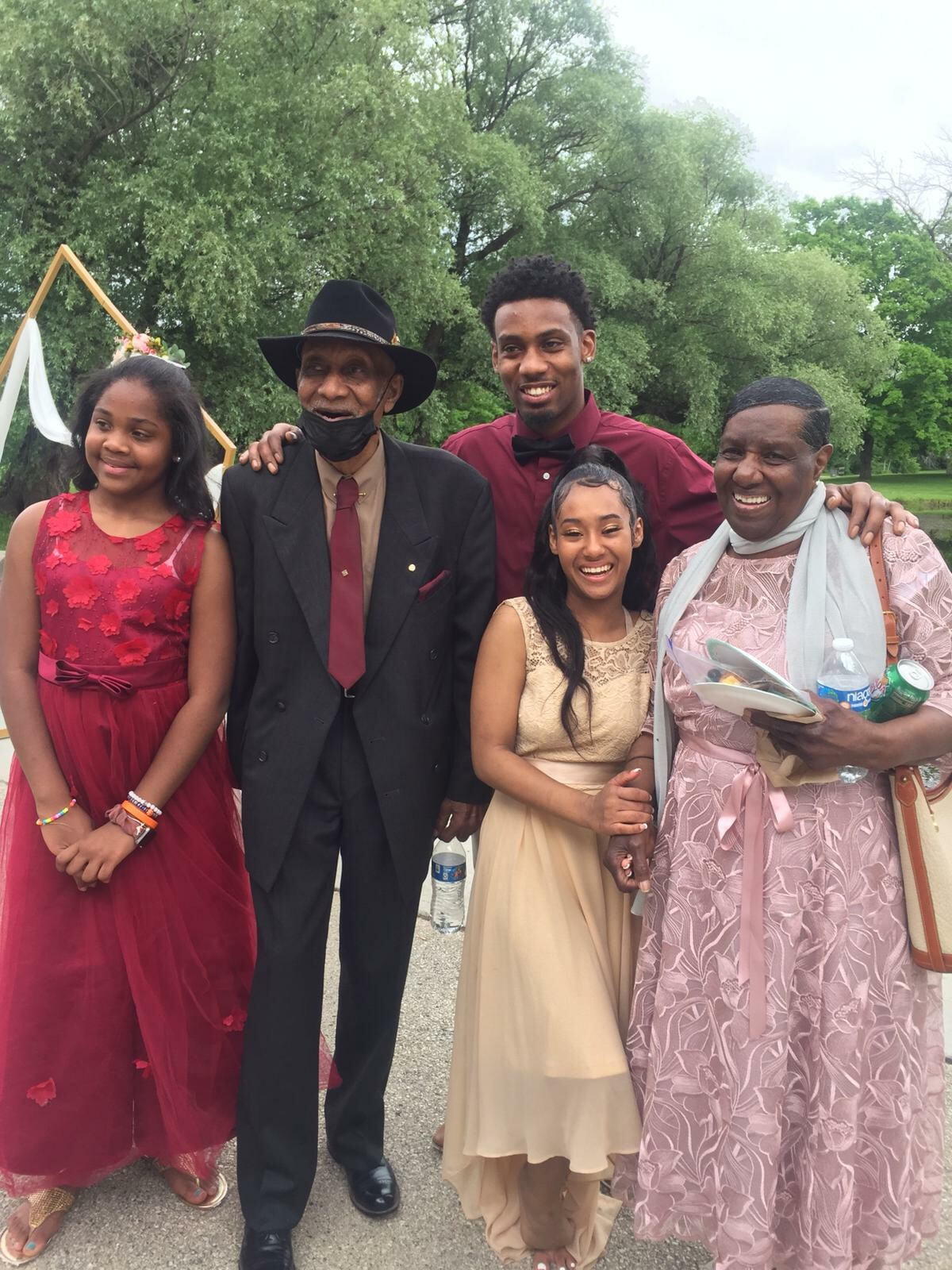 A multi-generational family posing together at an outdoor event, dressed in formal red, cream, and pink outfits, smiling at the camera.