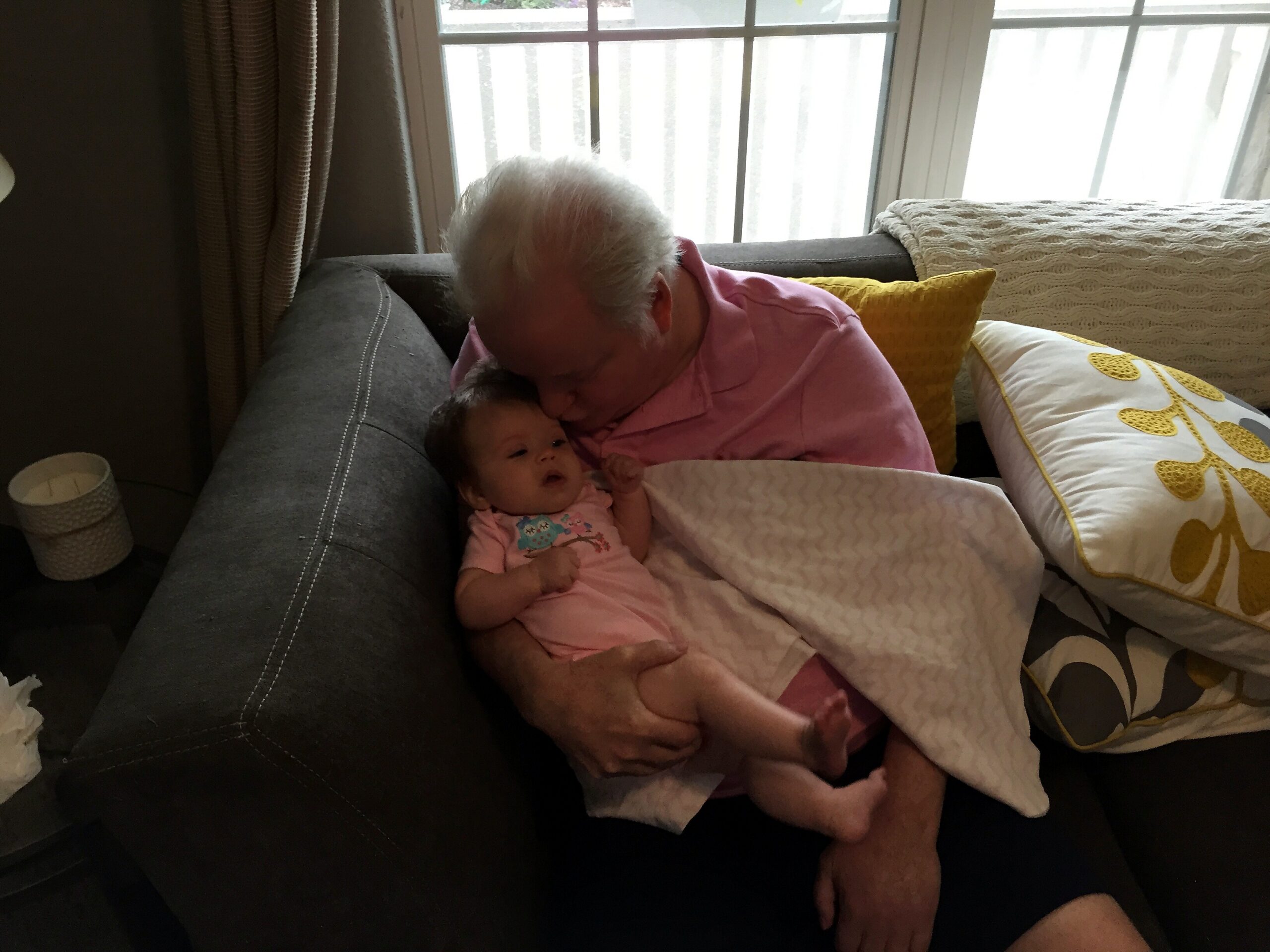 Elderly person with white hair wearing a pink shirt cradling a newborn baby on a gray sofa, near a window with yellow and white pillows nearby.