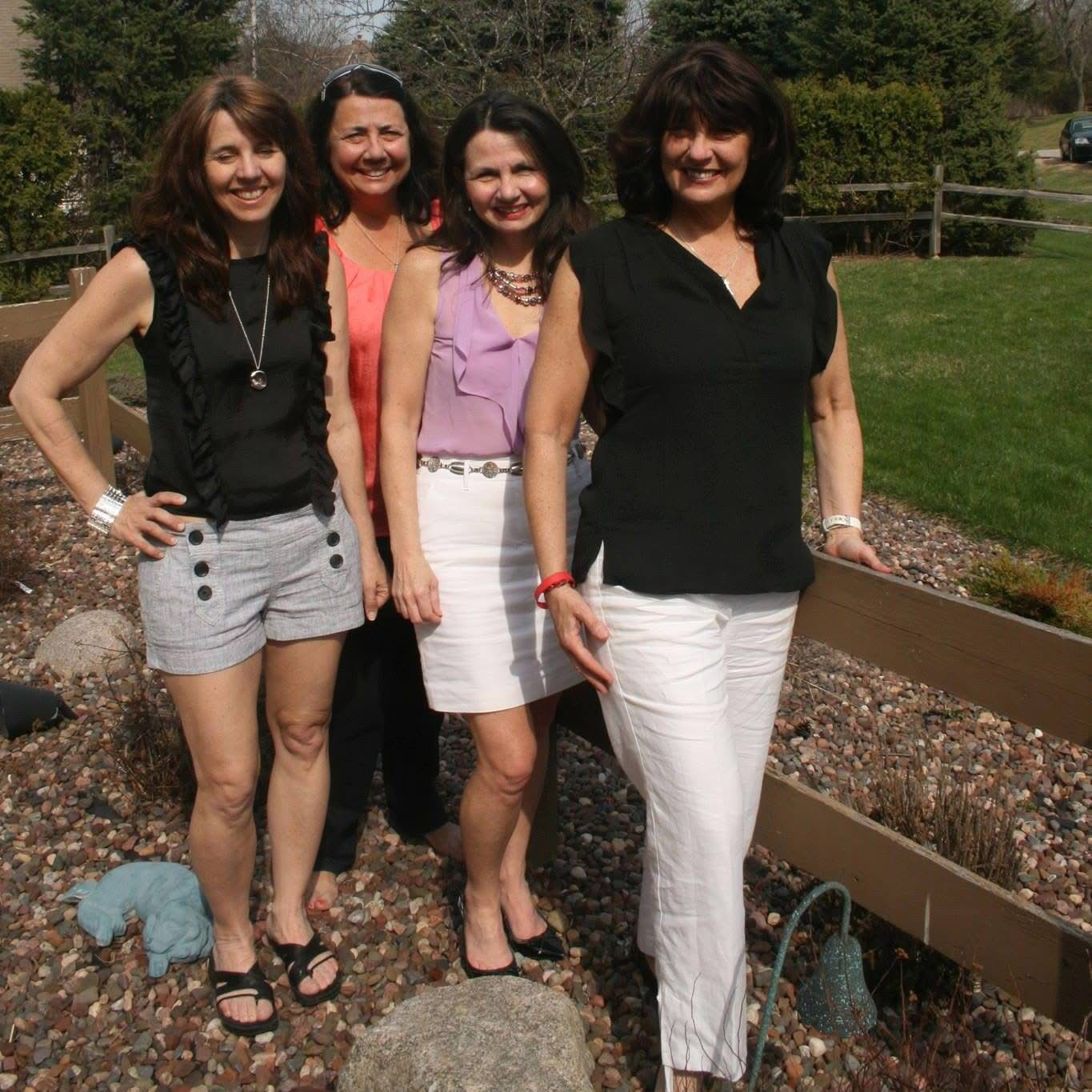 Four women posing together outside on a sunny day, smiling near a wooden fence and rocky ground area.
