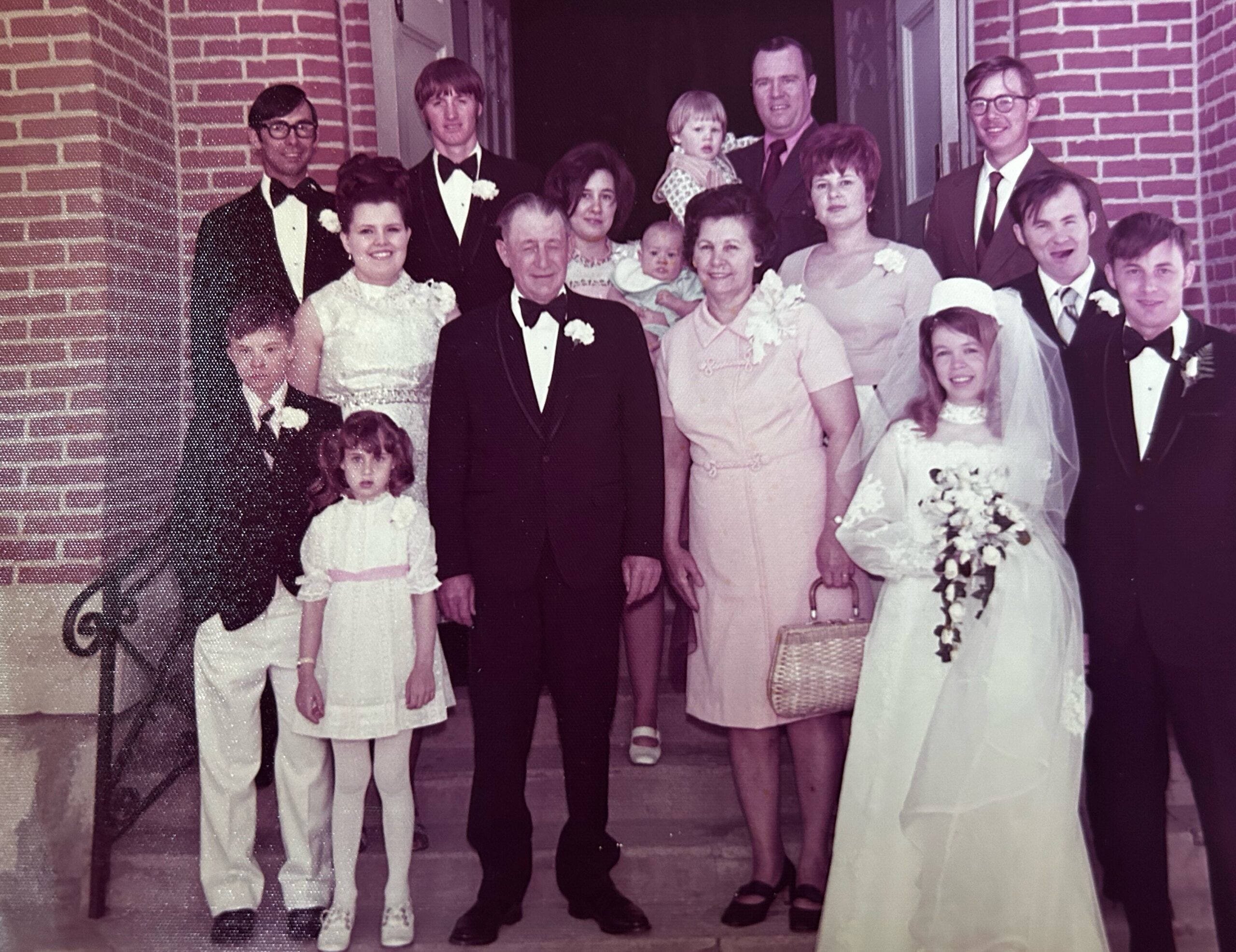 A large wedding party posing on steps outside a brick building, adults in tuxedos and dresses with a bride and groom at center.