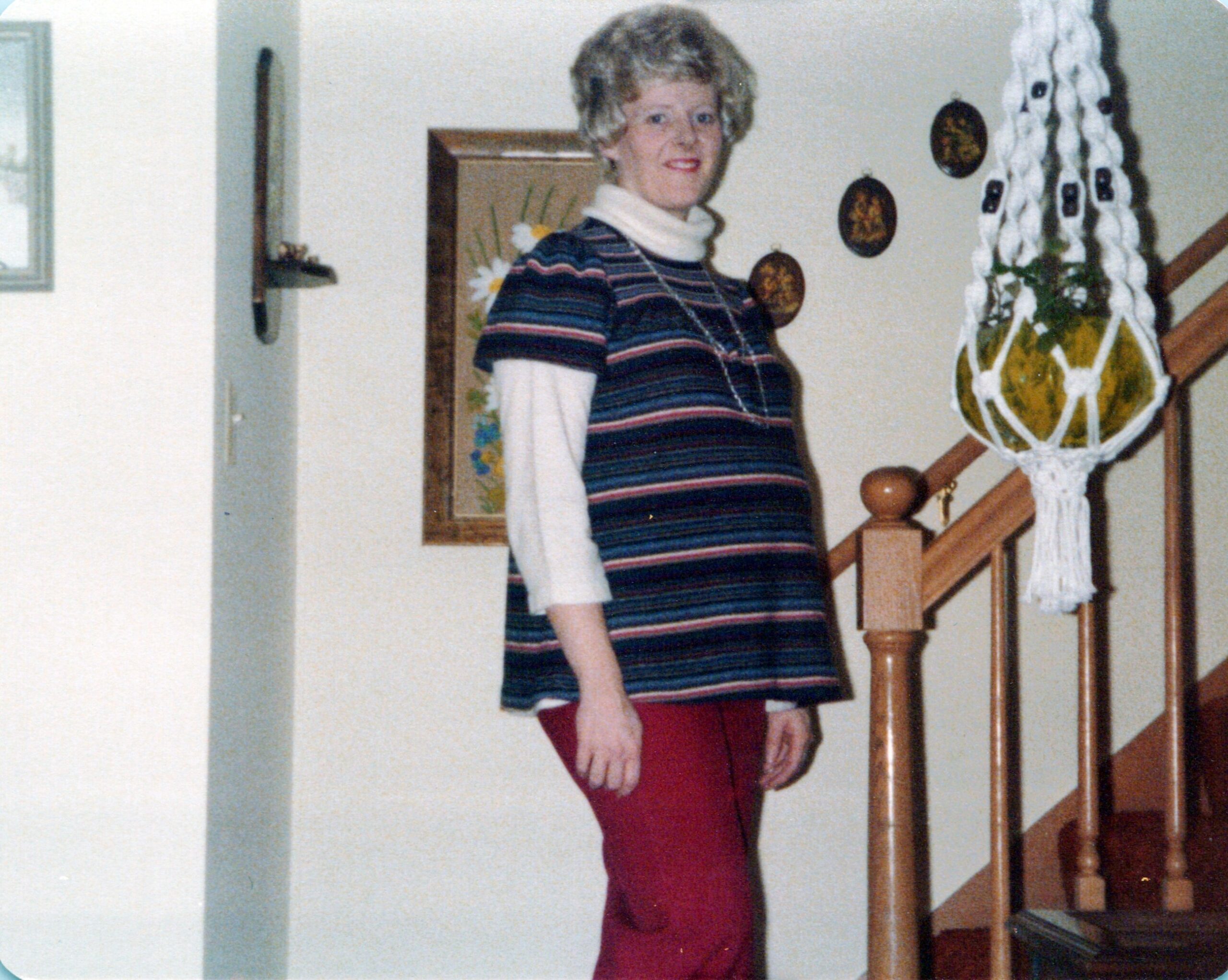 Woman standing on a wooden staircase indoors, wearing a striped knit top over a white turtleneck and red pants, smiling at the camera.