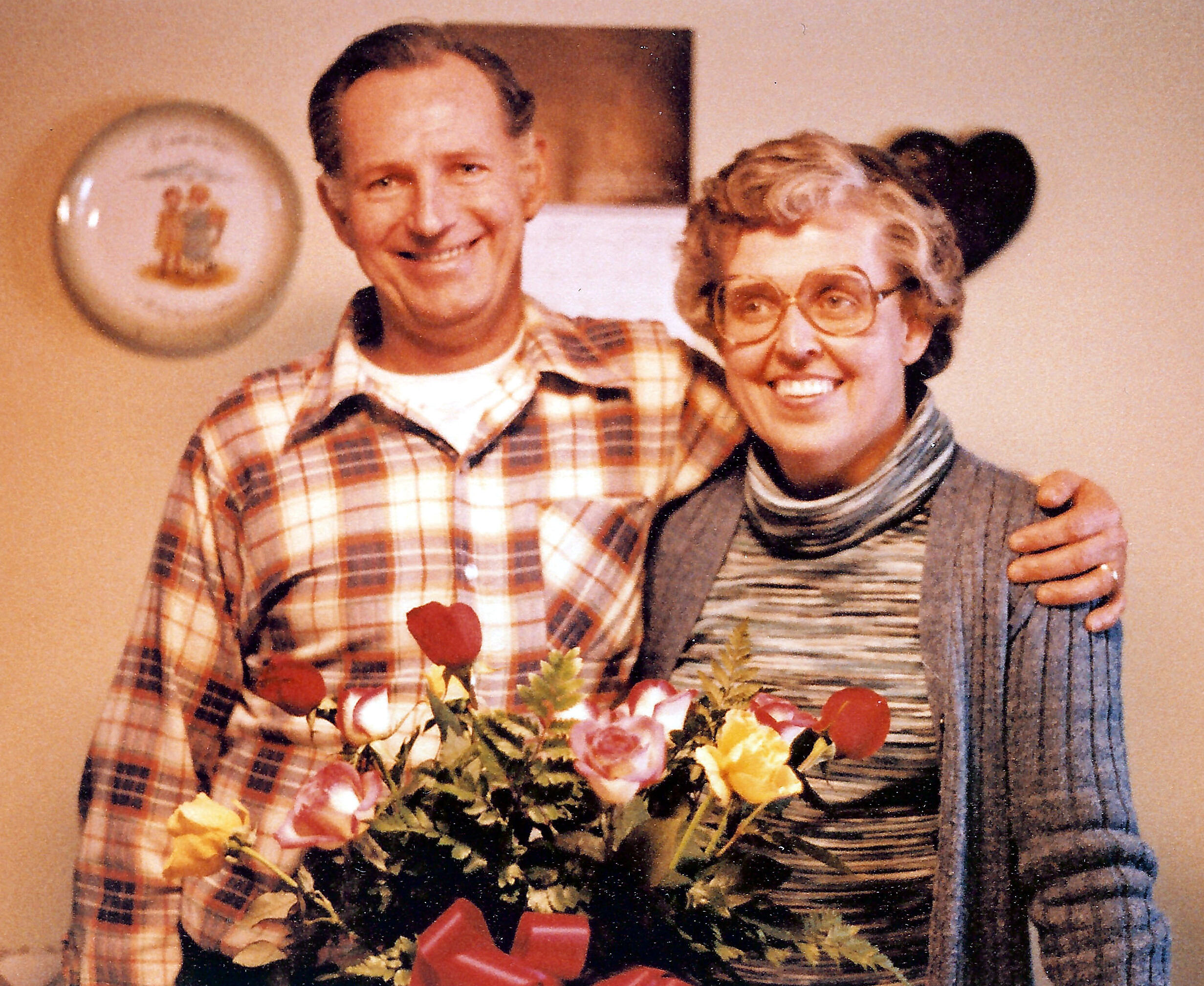 Older couple posing indoors, smiling at the camera with a bouquet of colorful flowers in front of them.