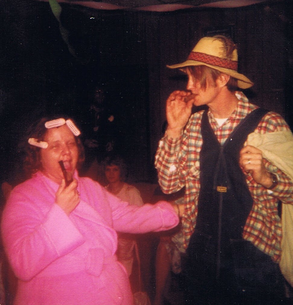 Two people in a playful scene: a woman in a pink robe with hair curlers eating a chocolate bar while a man in a straw hat and plaid shirt pretends to inspect him, on a stage or party setting.