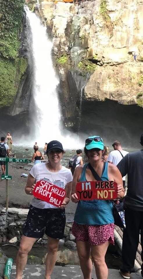Two women stand at a waterfall holding red handmade signs, smiling at the camera; one sign reads 'SAY HELLO FROM BALL' and the other 'I'M HERE YOU NOT'.
