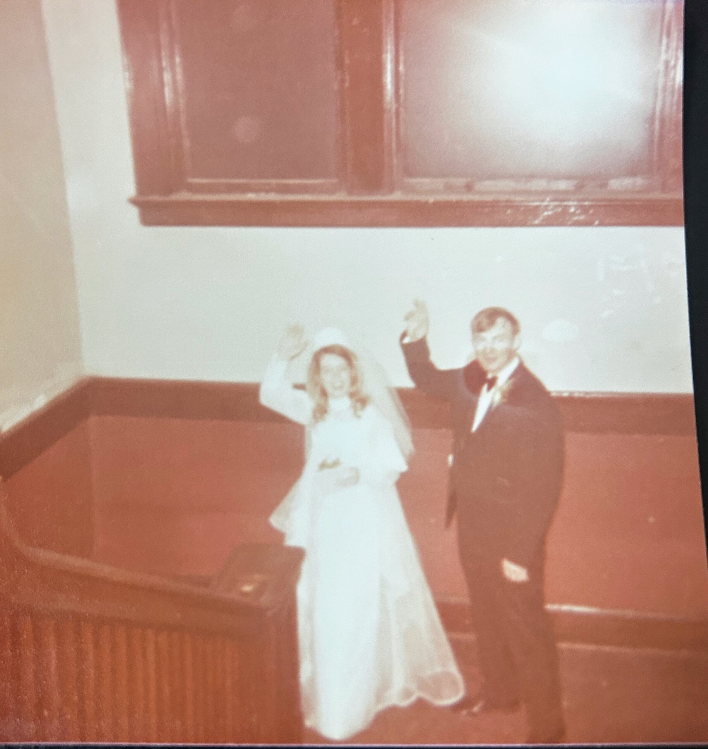 Bride in a white wedding gown and a man in a dark suit stand together, smiling, with other people nearby in an indoor setting.