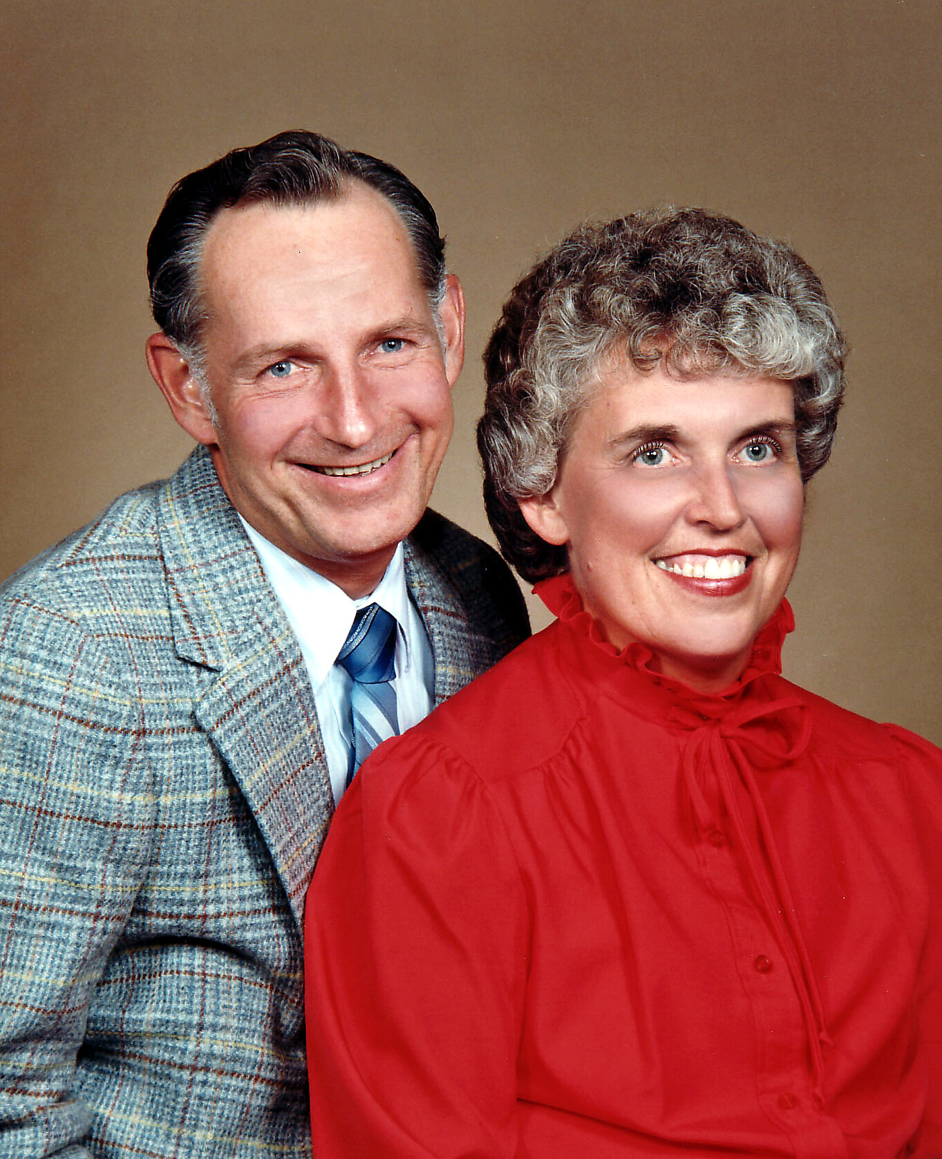 Smiling man in a gray plaid blazer and blue tie beside a smiling woman in a red blouse, studio portrait.