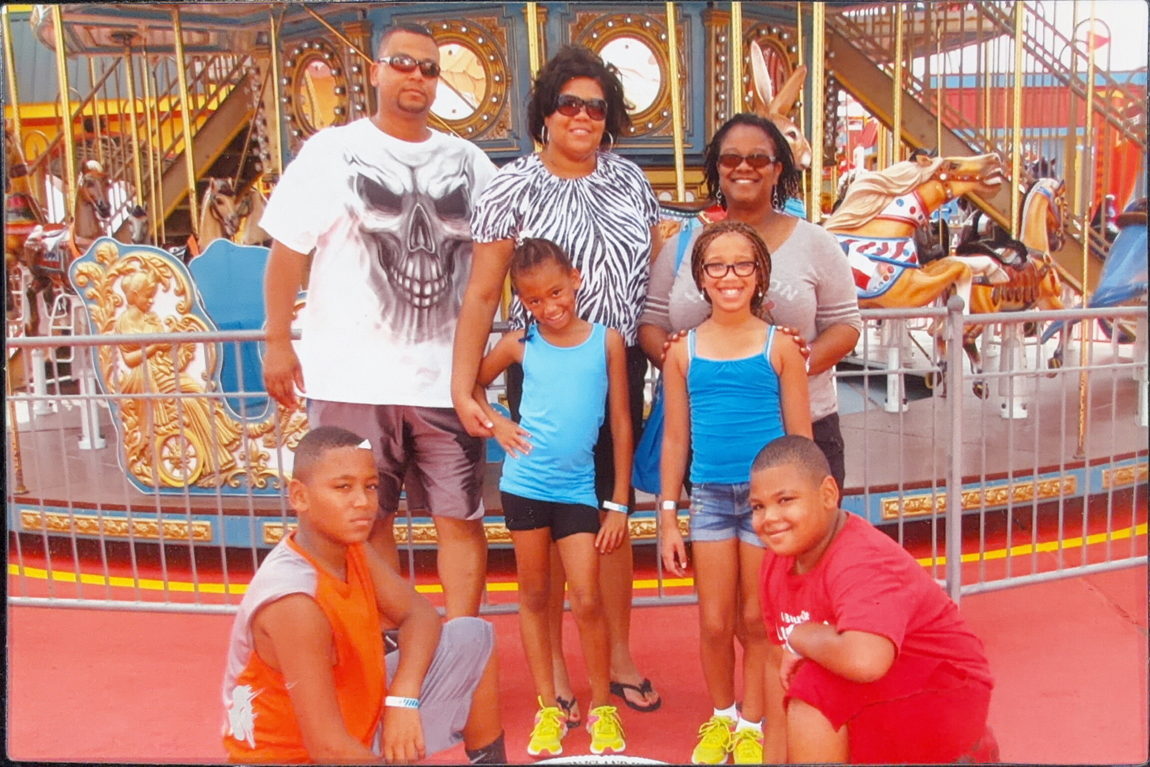 Group of adults and children posing in front of a bright carousel with painted horses at an amusement park.