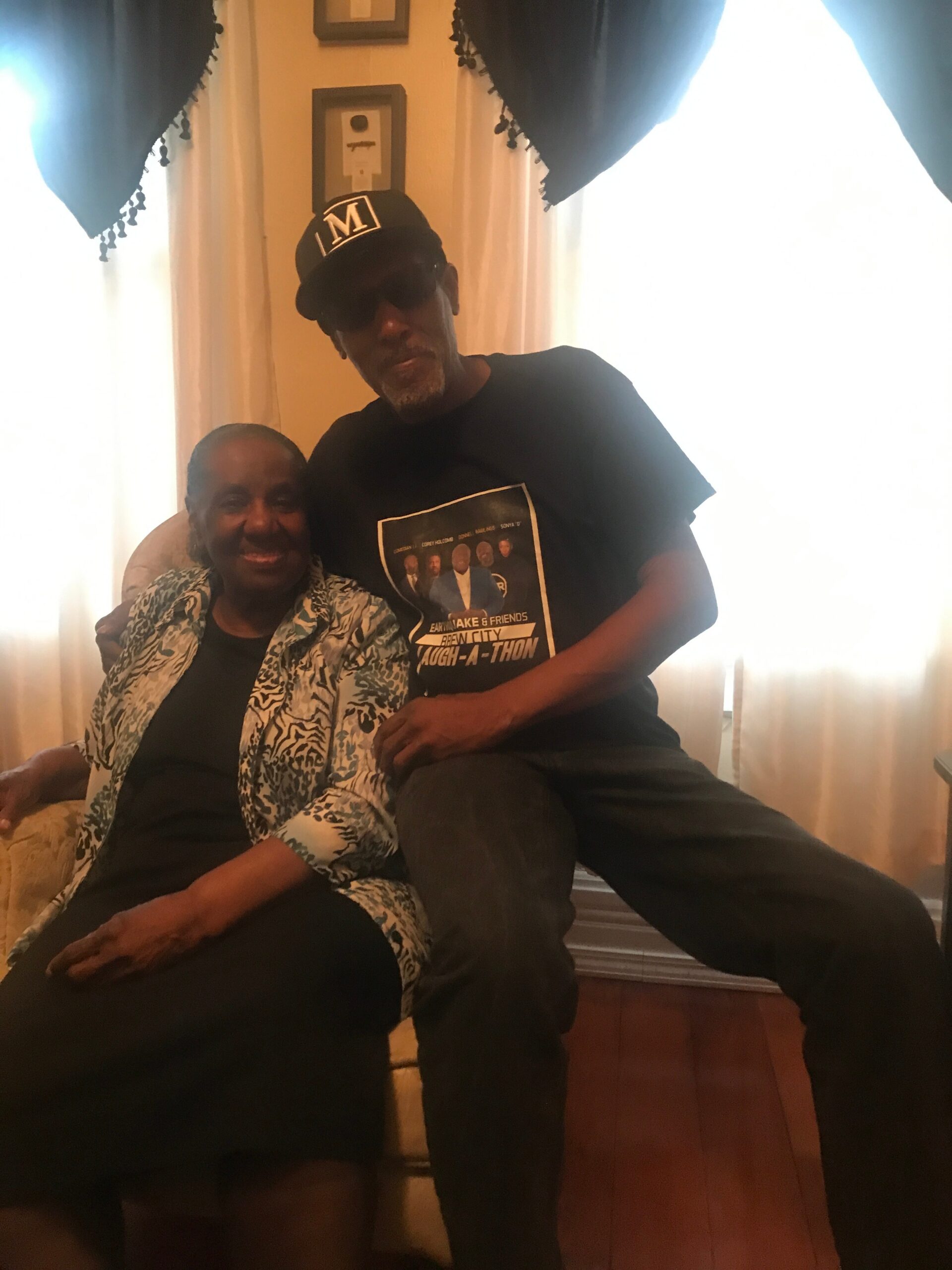 Elderly woman seated beside a man who has his arm around her in a living room; both smiling at the camera, man wearing a cap with 'LM' and a graphic tee.