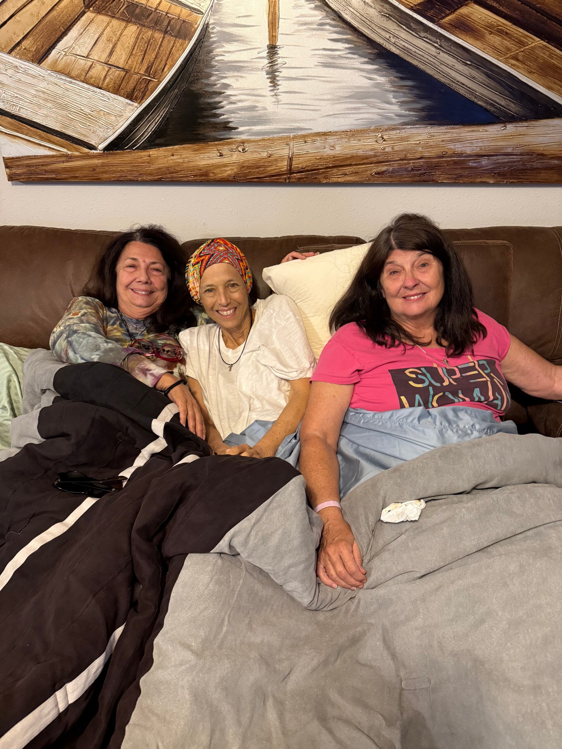 Three women sit close together on a sofa in a living room, smiling, with a boat painting on the wall behind them.