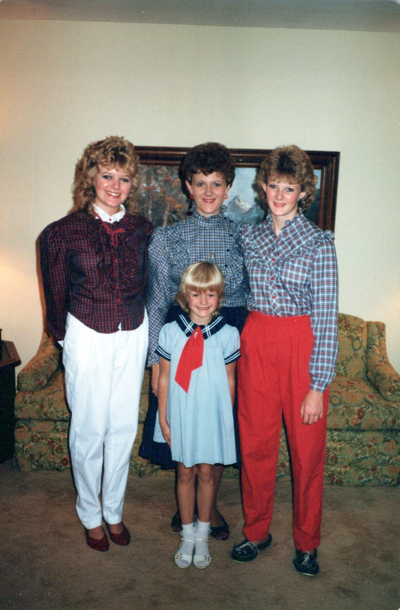 Four girls standing together in a living room, posing for a family photo in front of a floral couch and painting; the older girls wear plaid tops and red/white bottoms, while the youngest girl wears a light blue sailor-style dress with a red scarf.