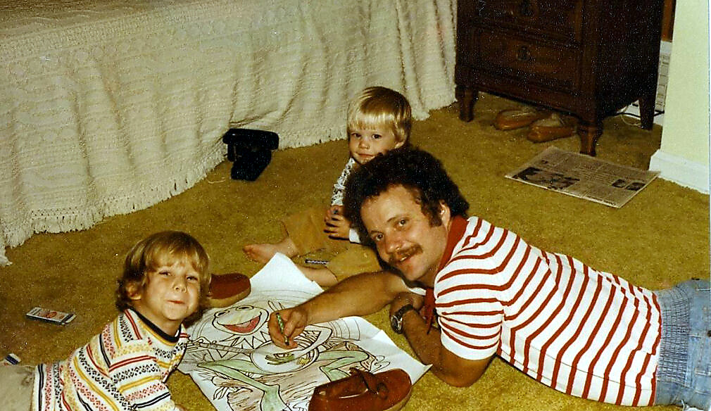 Man with a mustache and striped shirt lies on a beige carpet, coloring a large drawing with two young children nearby.