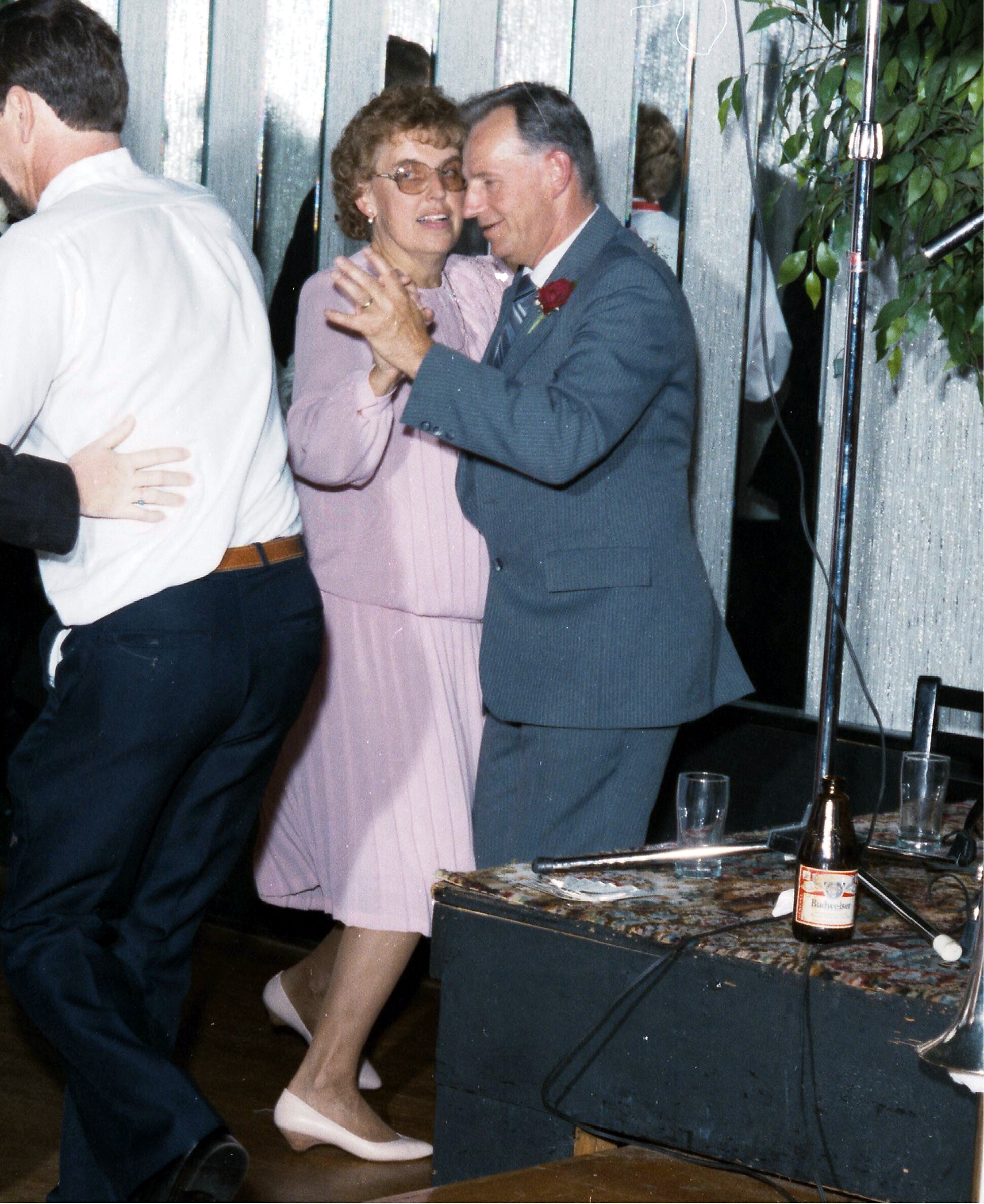 Older couple sharing a slow dance at a celebration; woman in pink dress and glasses, man in a gray suit with a red boutonniere.