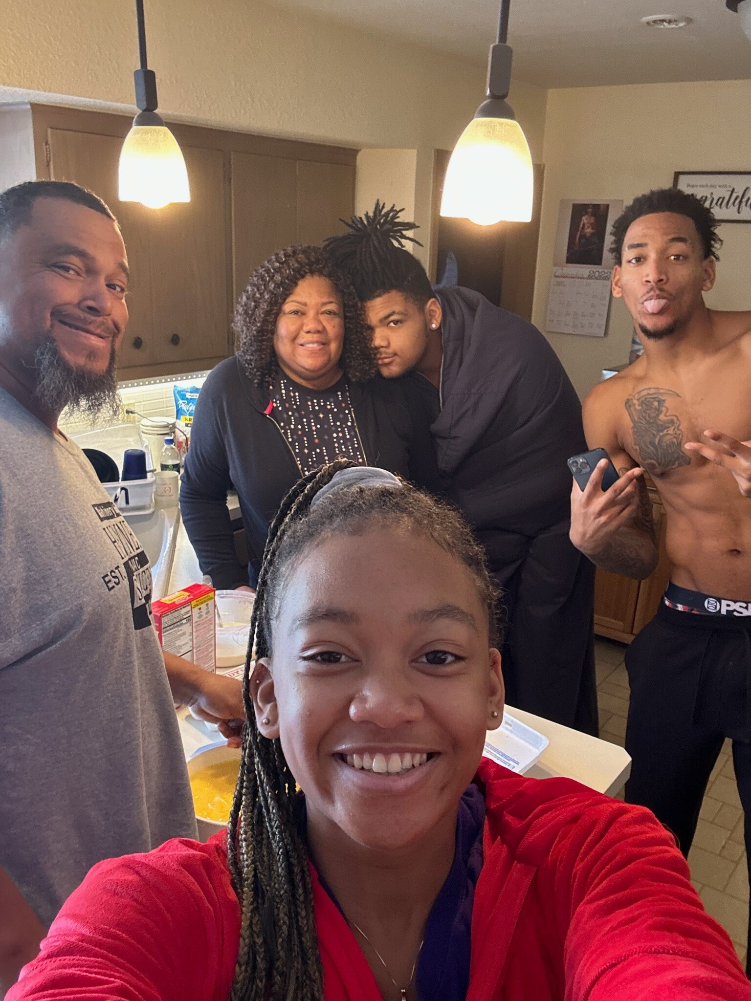 A group selfie in a kitchen: smiling woman in the center, teenagers and adults around her, with a shirtless man on the right sticking out his tongue.