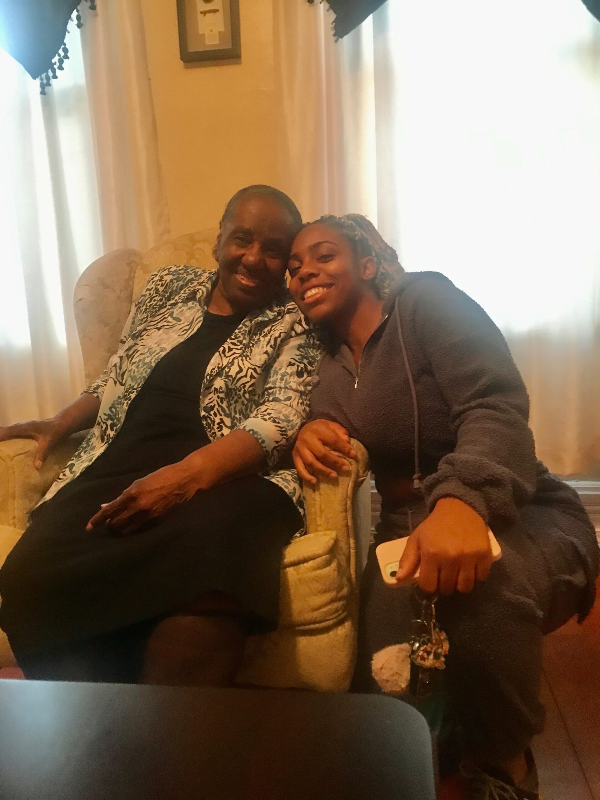 Two women sit close together on a beige armchair, smiling at the camera; older woman wears a patterned jacket, younger woman in a gray hoodie grip hands.