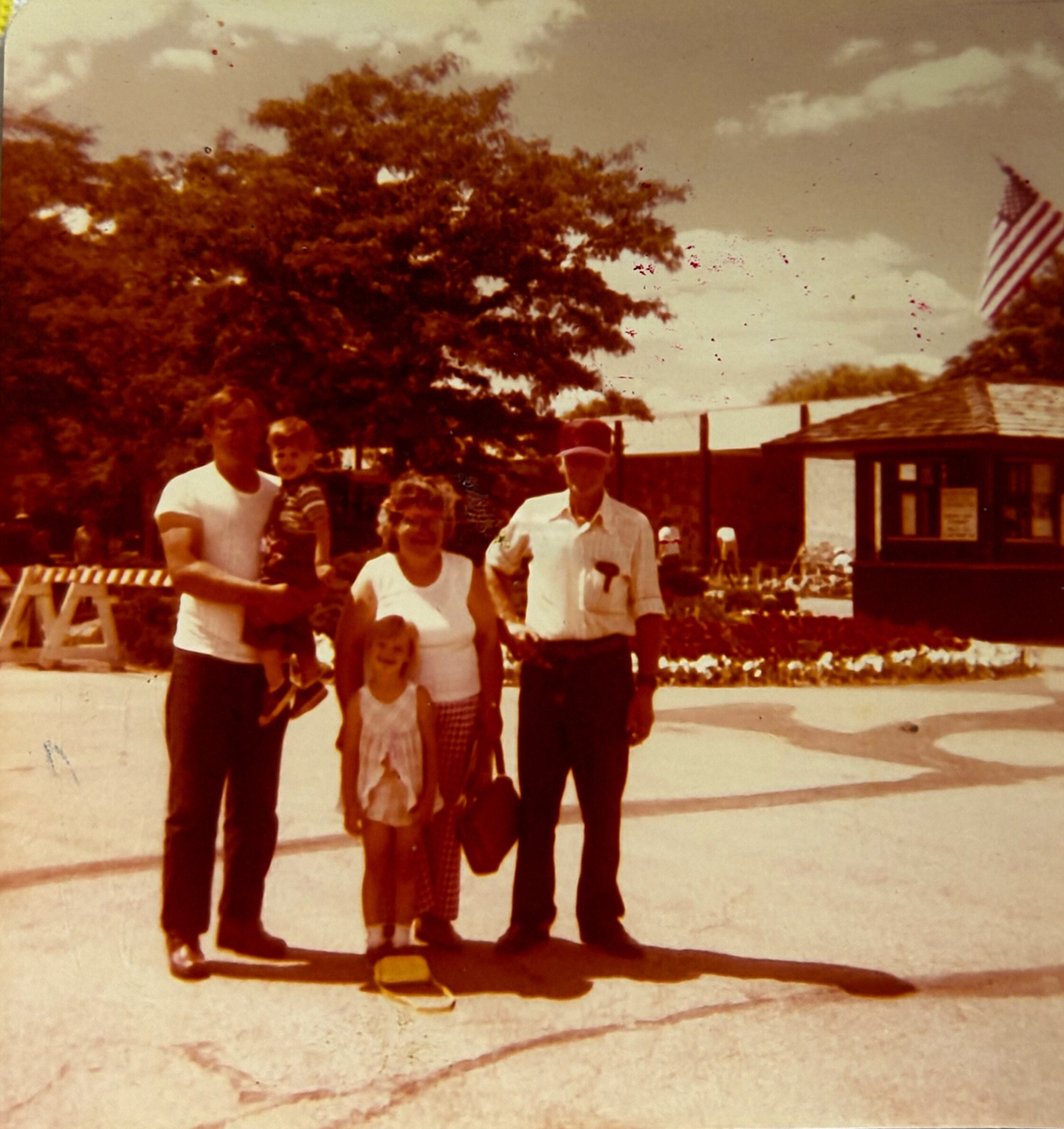 A family group posing outdoors in front of a fence, with a waving American flag in the background on the right.