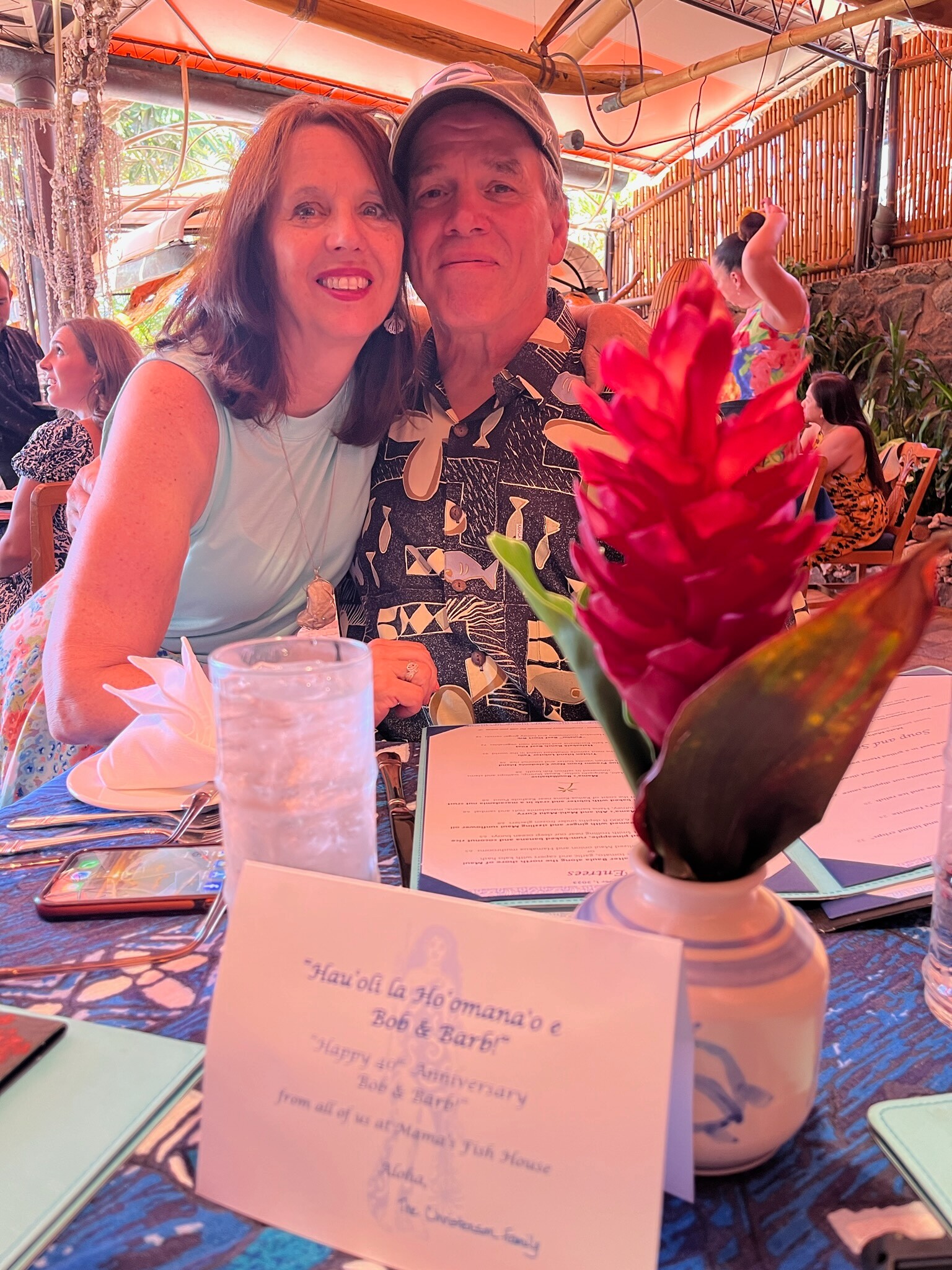Smiling couple seated at a dinner table with a bright red flower centerpiece and menus in a tropical restaurant setting.