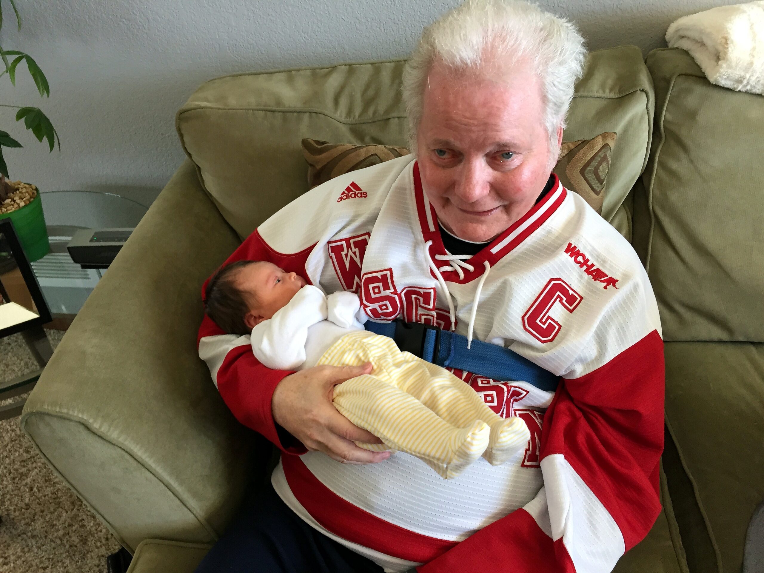 Senior man in a red-and-white jersey sits on a sofa, cradling a newborn wrapped in a yellow-striped blanket on his lap of a cozy living room setting.