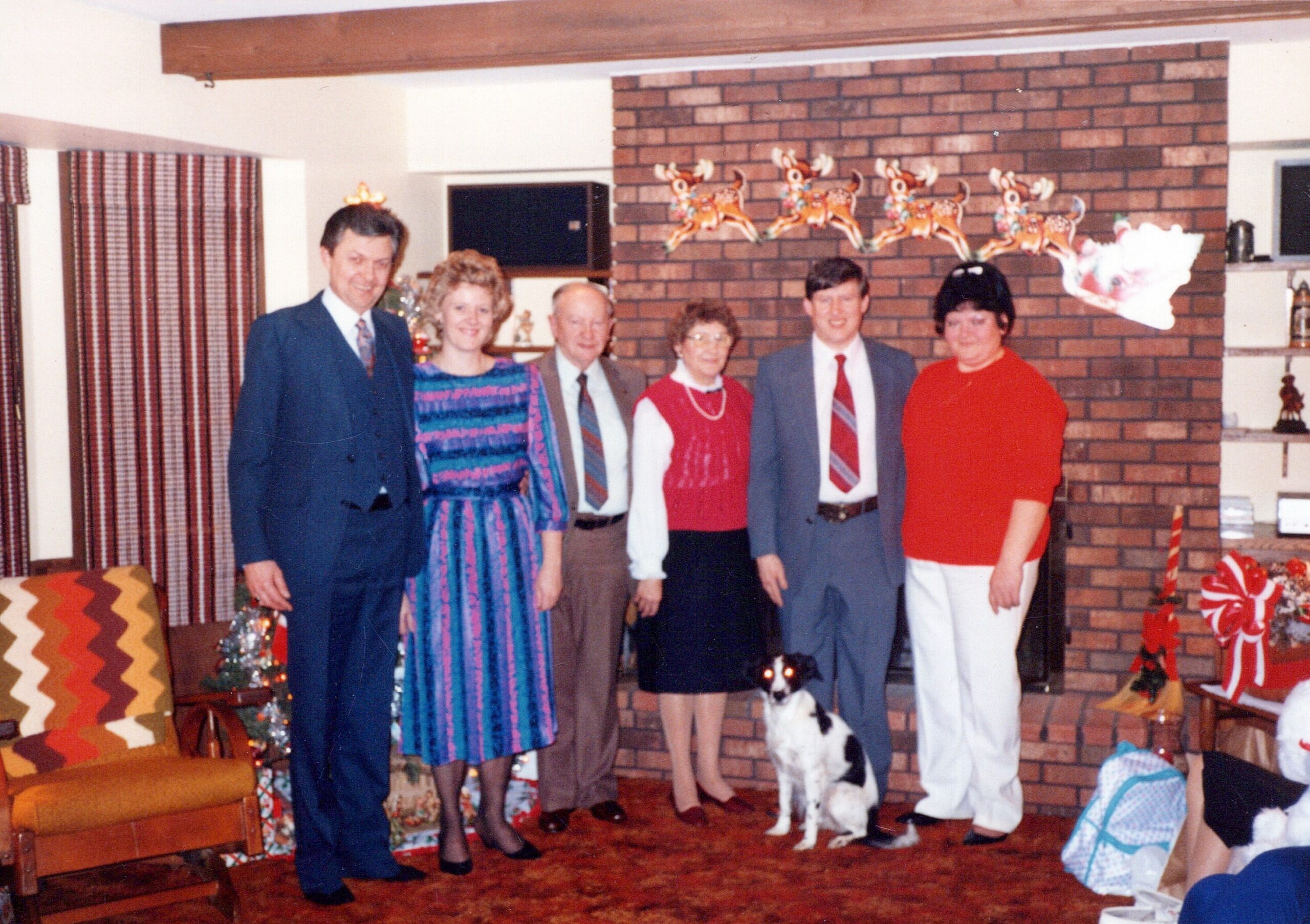 Group of adults (and a small dog) posing for a Christmas photo in a living room with a brick fireplace, stockings, and wrapped gifts in view.
