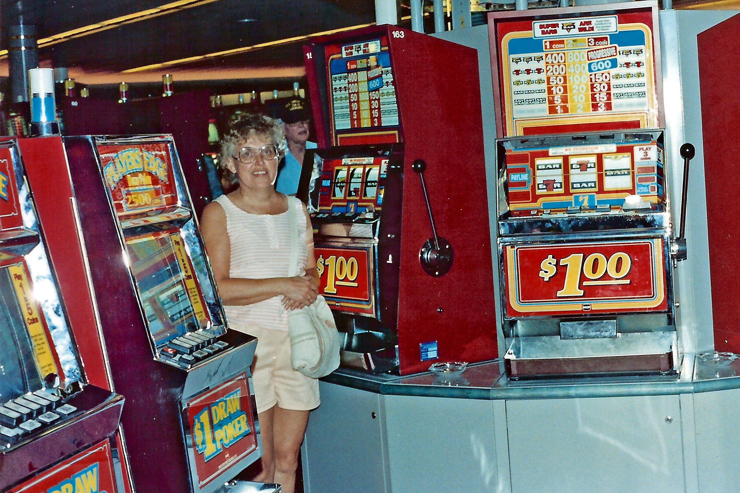 Woman in a light striped sleeveless top smiles while standing beside colorful vintage slot machines in a casino alley.