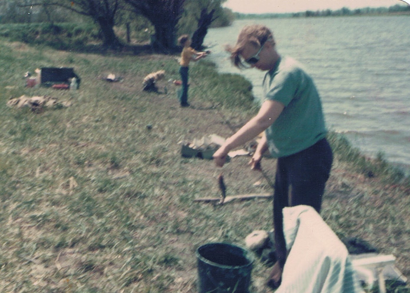 Man standing by a lakeshore, holding a small fish on a line with a rod, a bucket at his feet and a towel draped nearby as others fish in the background.