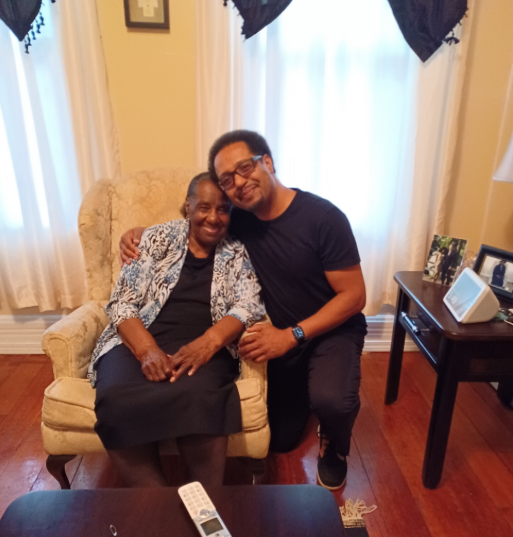 Smiling young man kneels beside an elderly woman in a beige armchair, arms around each other in a cozy living room.