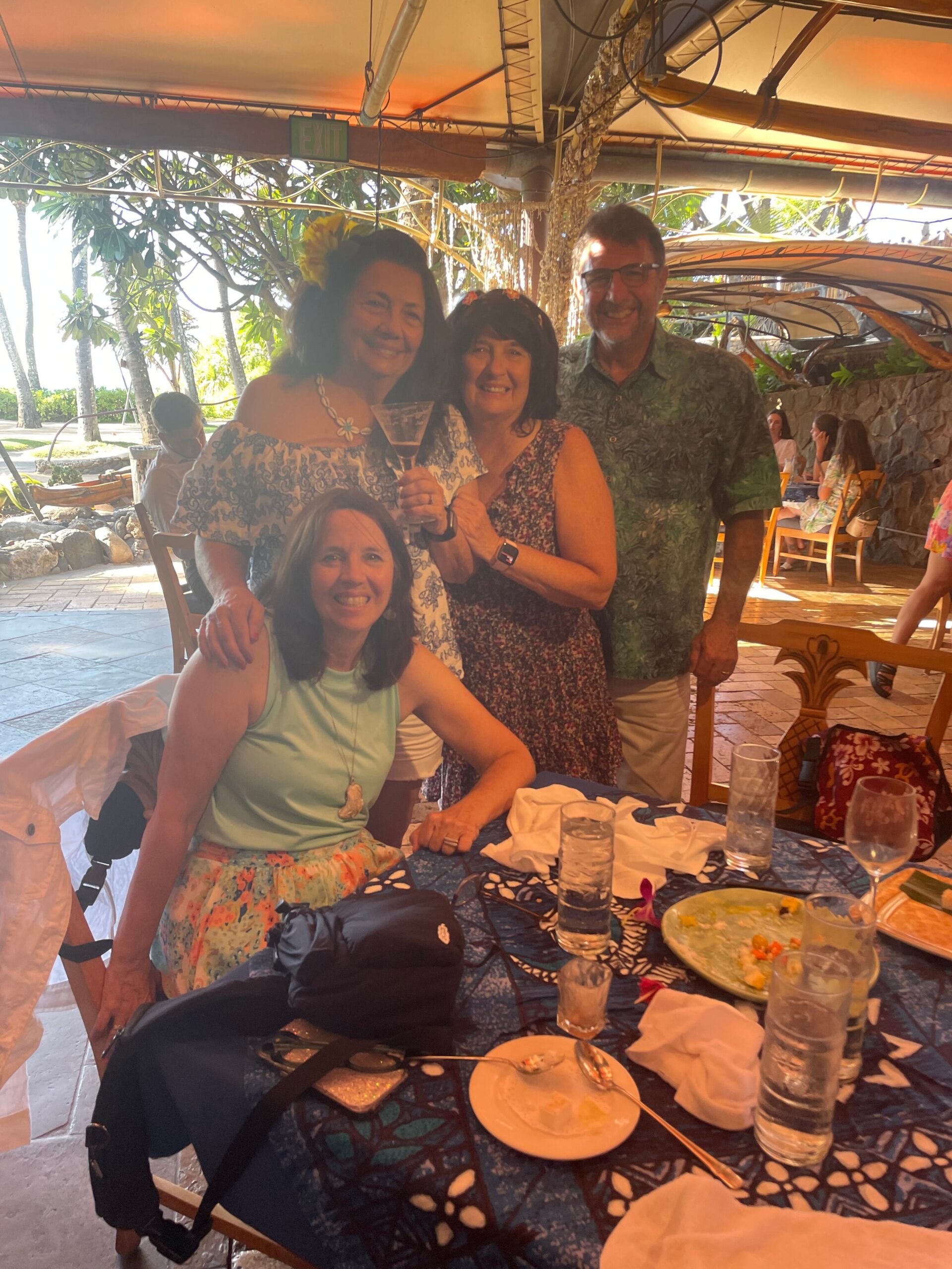 Group of four adults smiling at a tropical outdoor restaurant, posing with drinks near a decorated table and sunny greenery in the background, mid-meal scene.