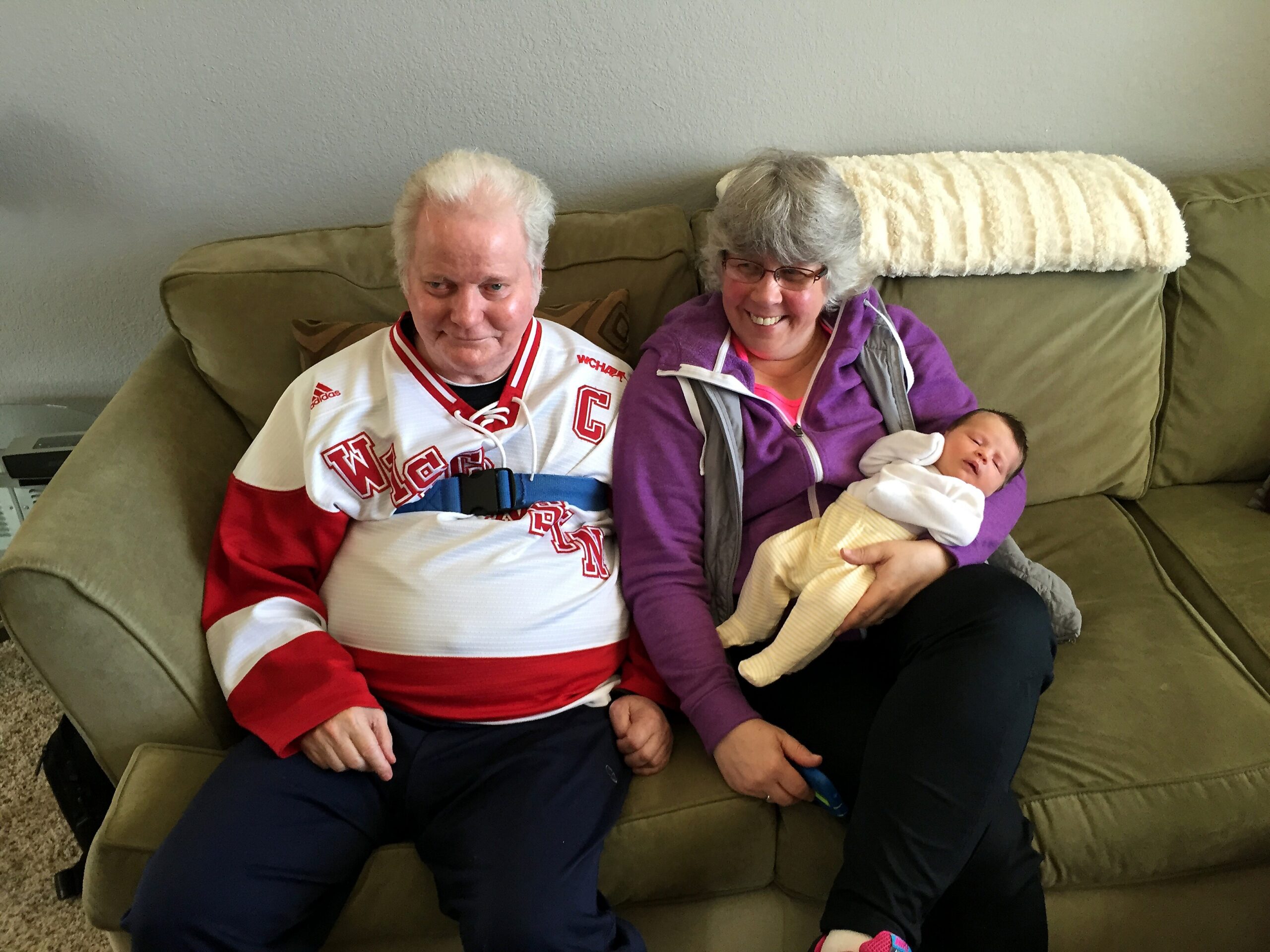 Two smiling adults sit close together on a green couch, with a newborn baby wrapped in a light blanket in the woman's arms.
