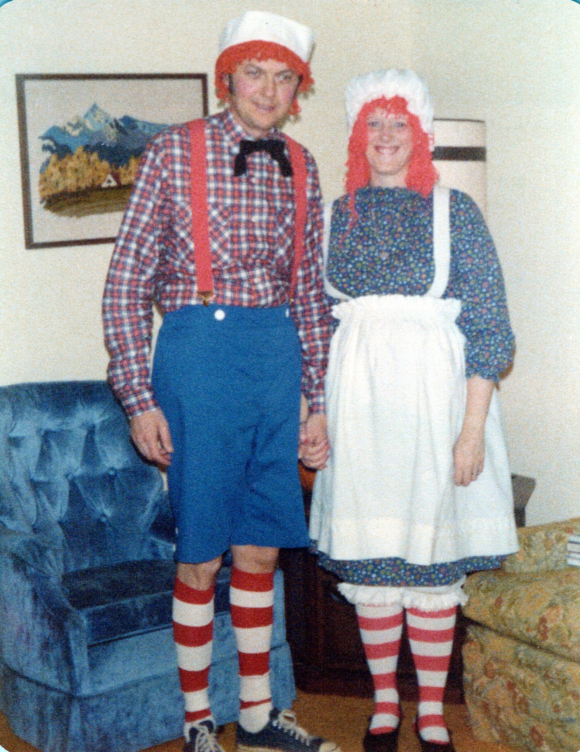 Two people in retro costumes stand together indoors: man with red suspenders, checkered shirt, blue shorts, bow tie and white hat; woman in floral dress with white apron and red wig with bonnet.