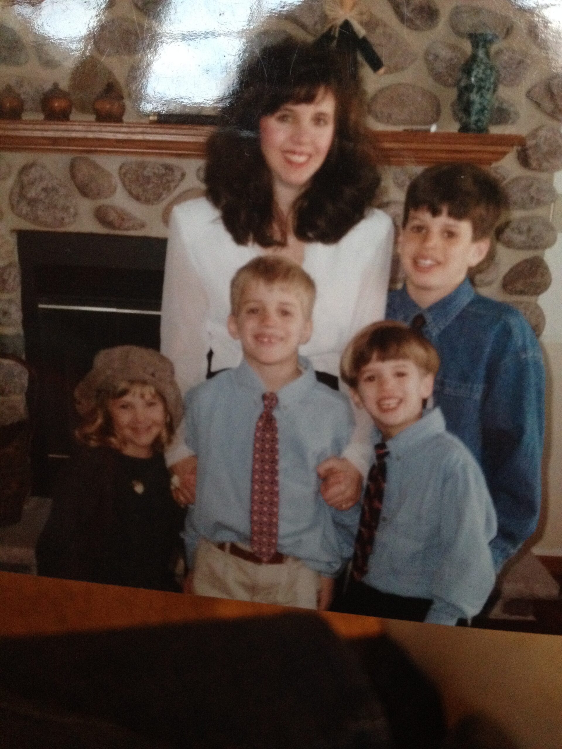 Family portrait in a living room: a woman with dark hair standing behind four children in blue shirts and ties in front of a stone fireplace.
