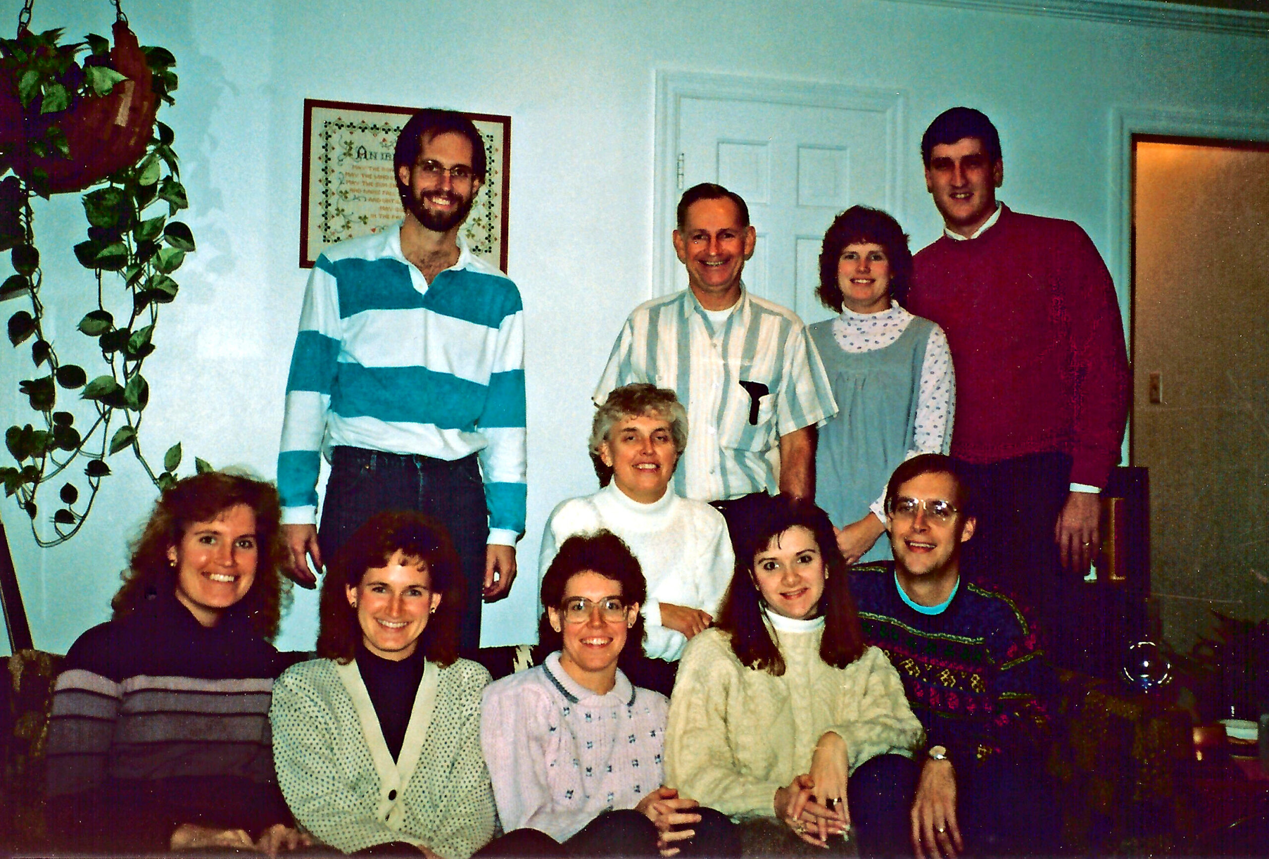 Group of smiling adults and teens posing indoors in a living room; light blue walls, a hanging plant on the left, and a framed picture behind them.