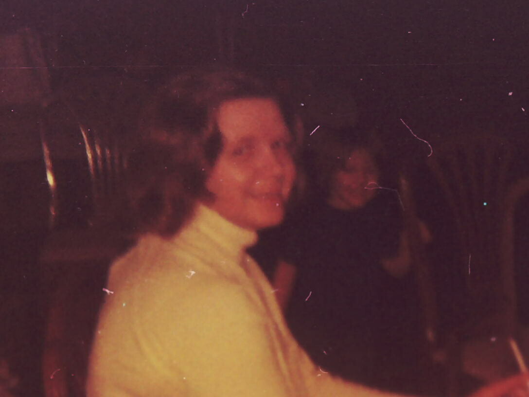 Woman with short curly hair wearing a light-colored blazer, smiling at the camera in a dim indoor setting; a darker-clad person sits in the background.