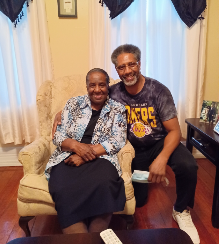 An elderly woman sits in a beige armchair, smiling, with a man kneeling beside her and his arm around her shoulders in a living-room setting.