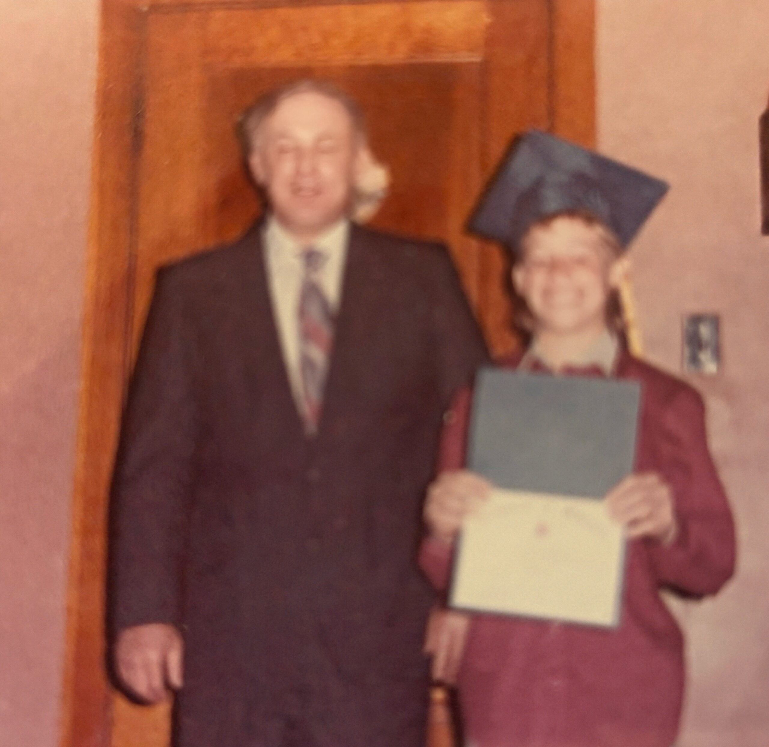 Man in a suit stands beside a graduate in a maroon gown and blue cap, holding a diploma or certificate in a hallway or doorway, both smiling (blurry photo).