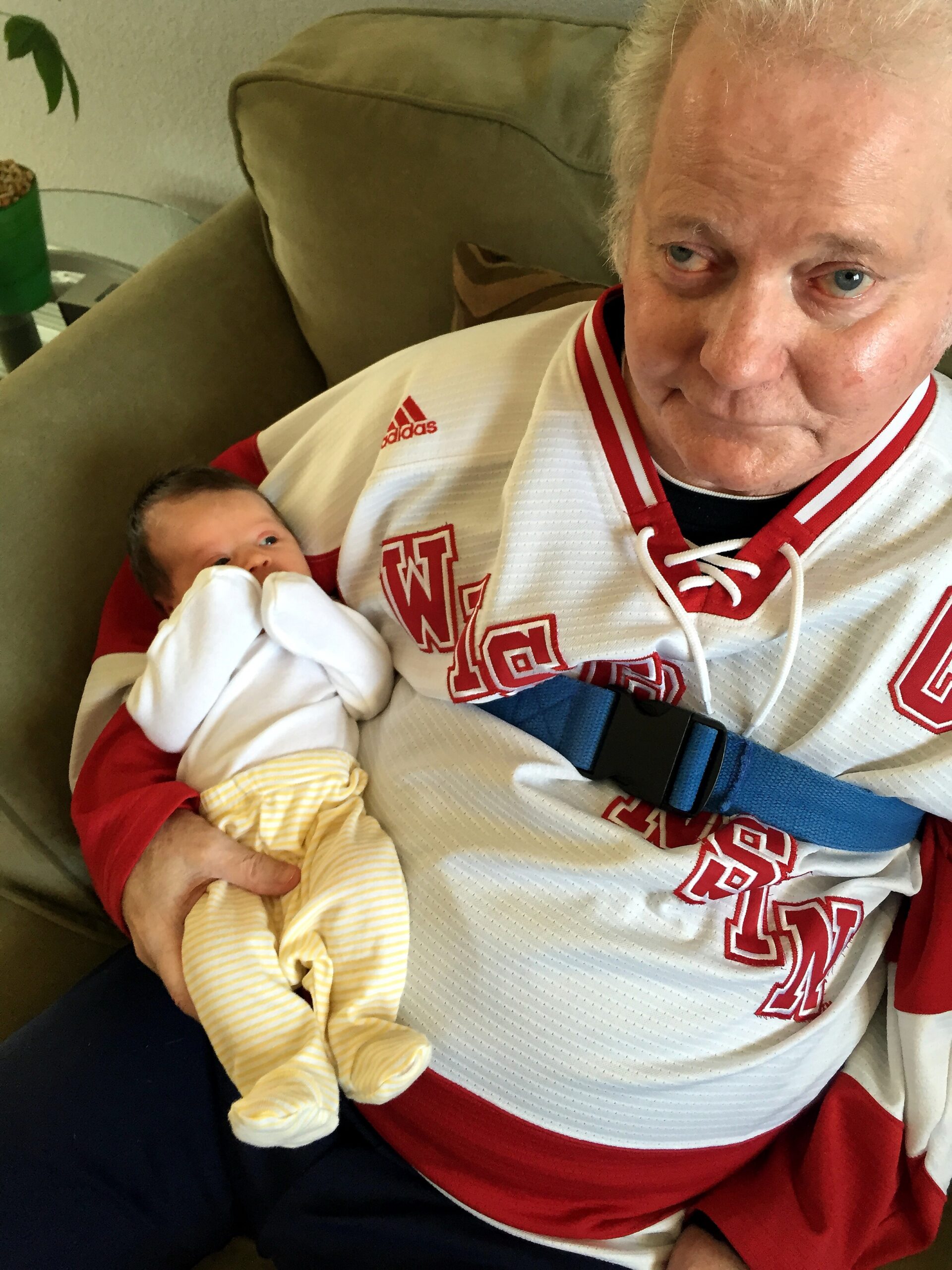 Elderly man in a white red athletic jersey cradling a newborn baby wrapped in a yellow striped onesie.
