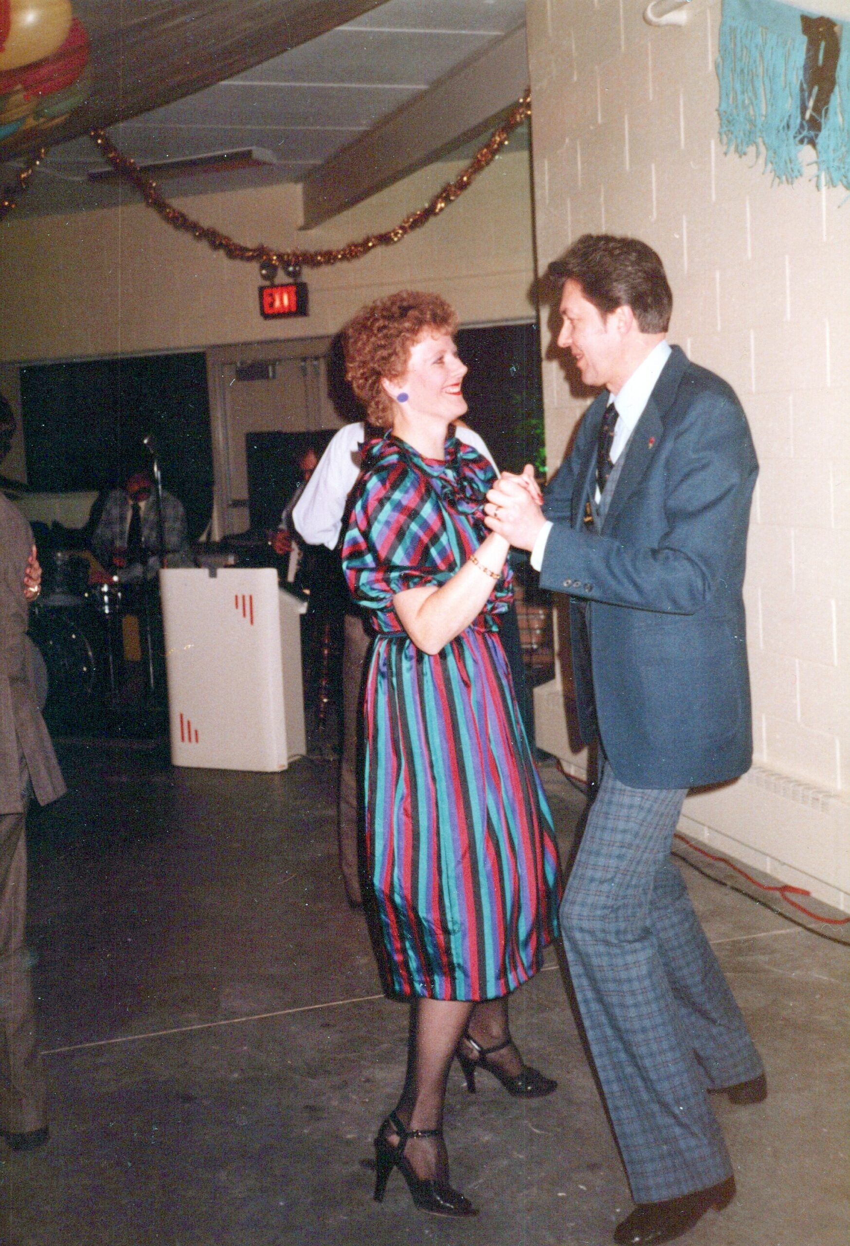 Couple dancing together at a party, smiling at each other in vintage outfits with colorful striped dress and checked pants under party decorations.