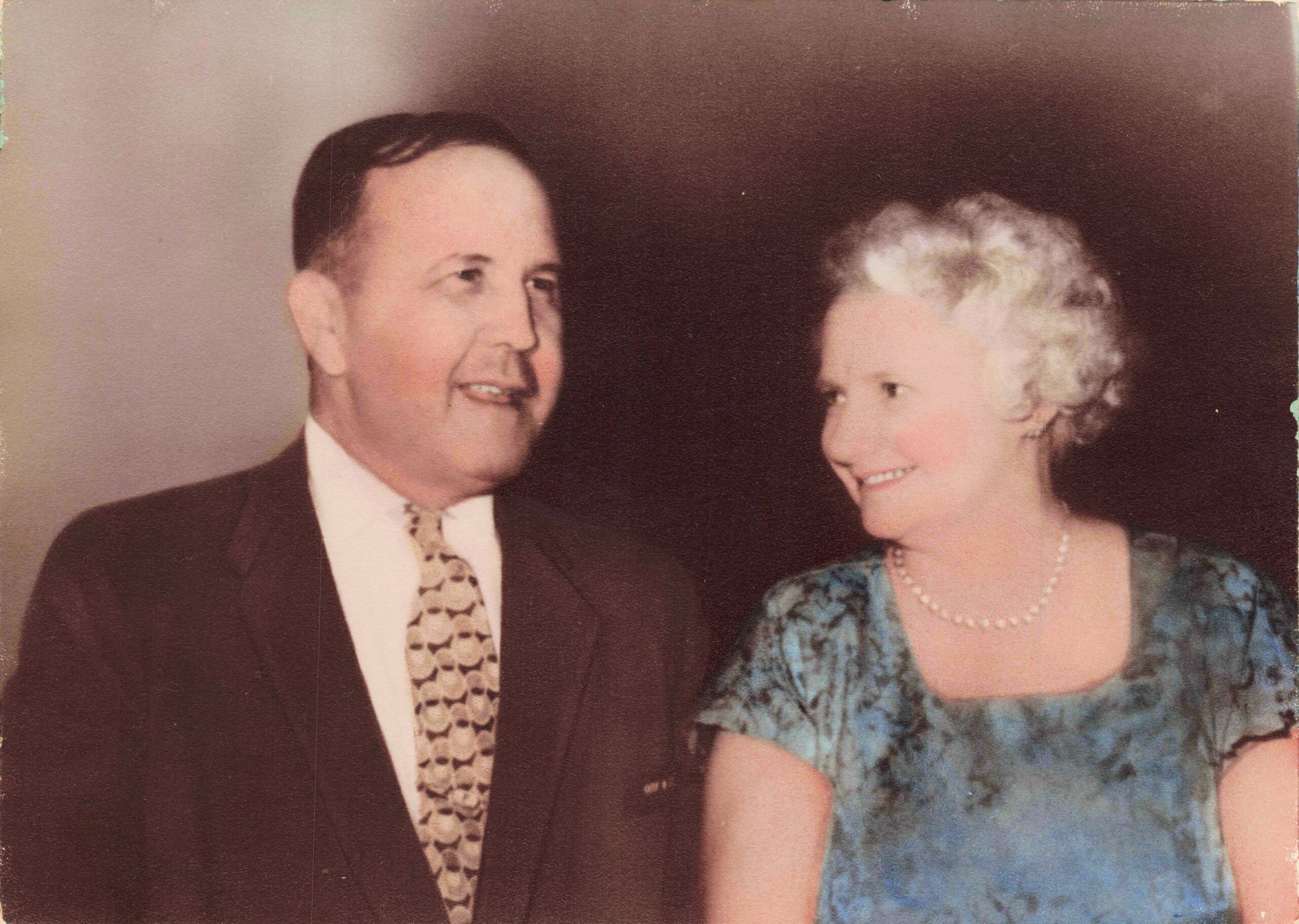 Smiling man in a dark suit and patterned tie stands beside a woman in a blue lace dress with a pearl necklace, both looking at each other in a formal portrait.