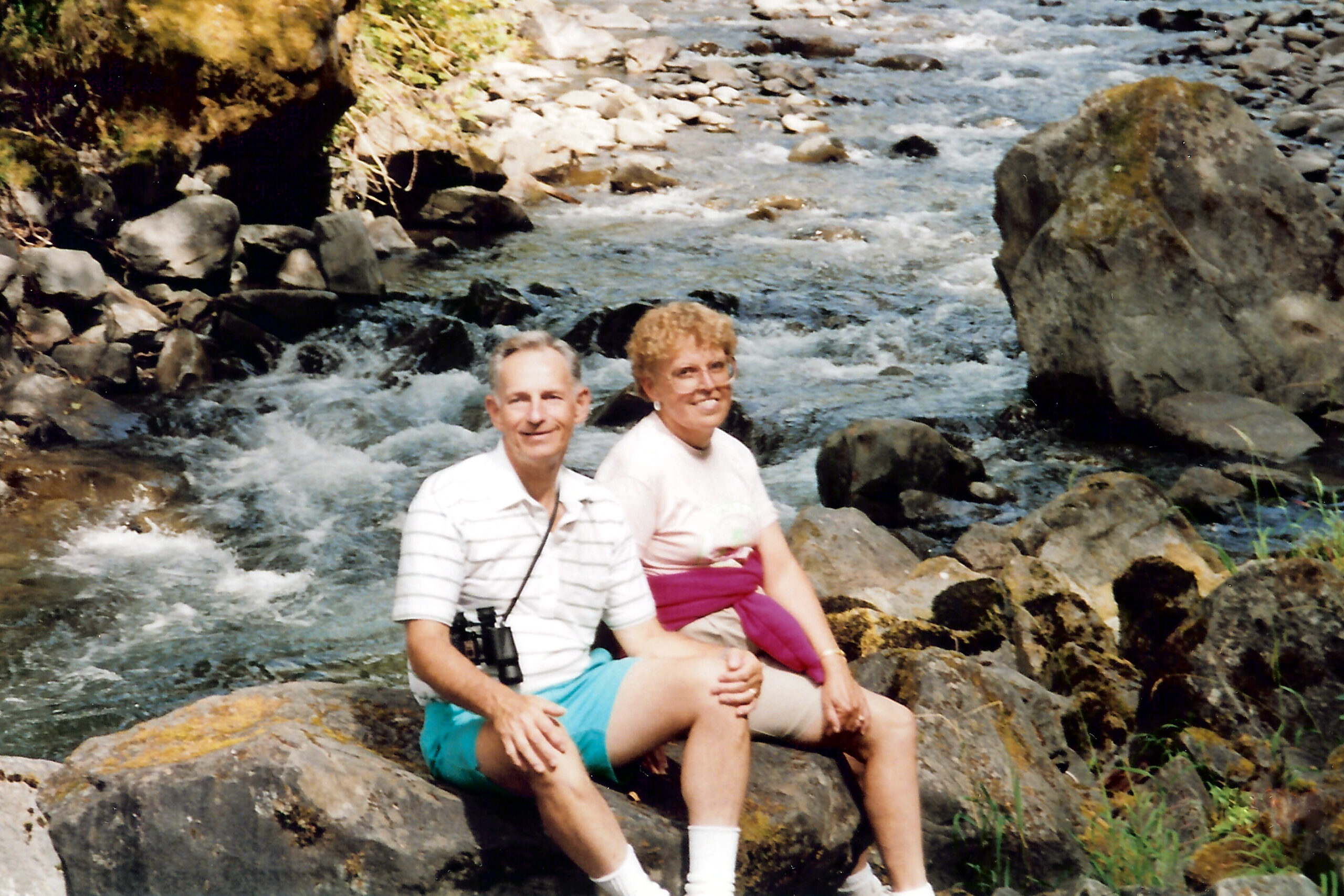 Older couple sitting on rocks beside a fast-flowing river in a rocky outdoor setting.